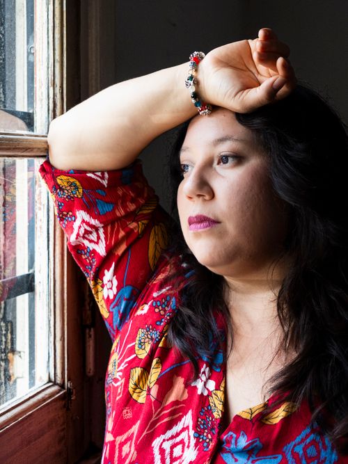 Free A woman in a colorful dress gazes thoughtfully out a window, hand resting on her forehead. Stock Photo