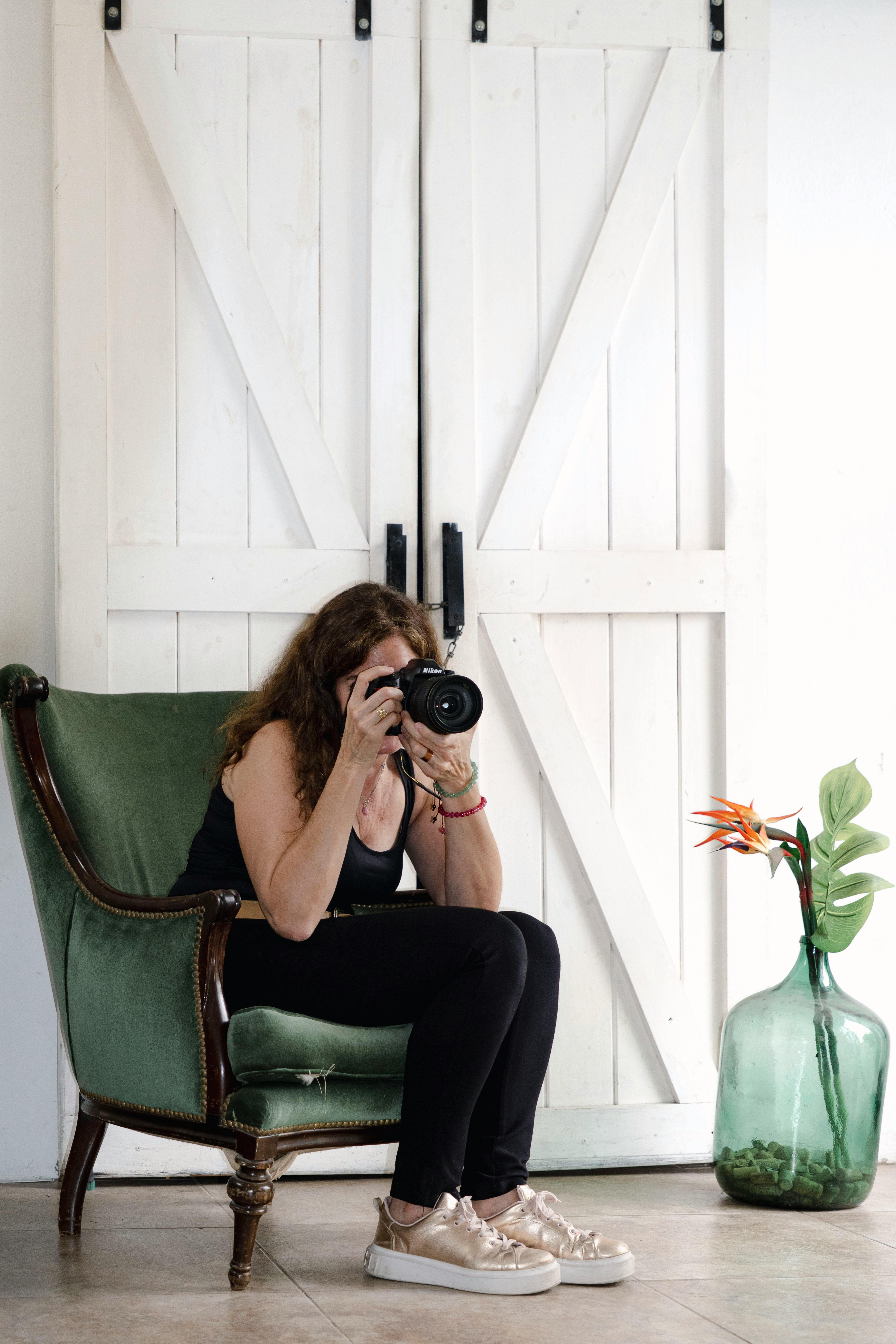 A female photographer sitting on a chair in a studio with rustic white doors and a decorative plant.