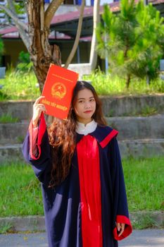 Young Vietnamese woman in graduation attire holding diploma outdoors.