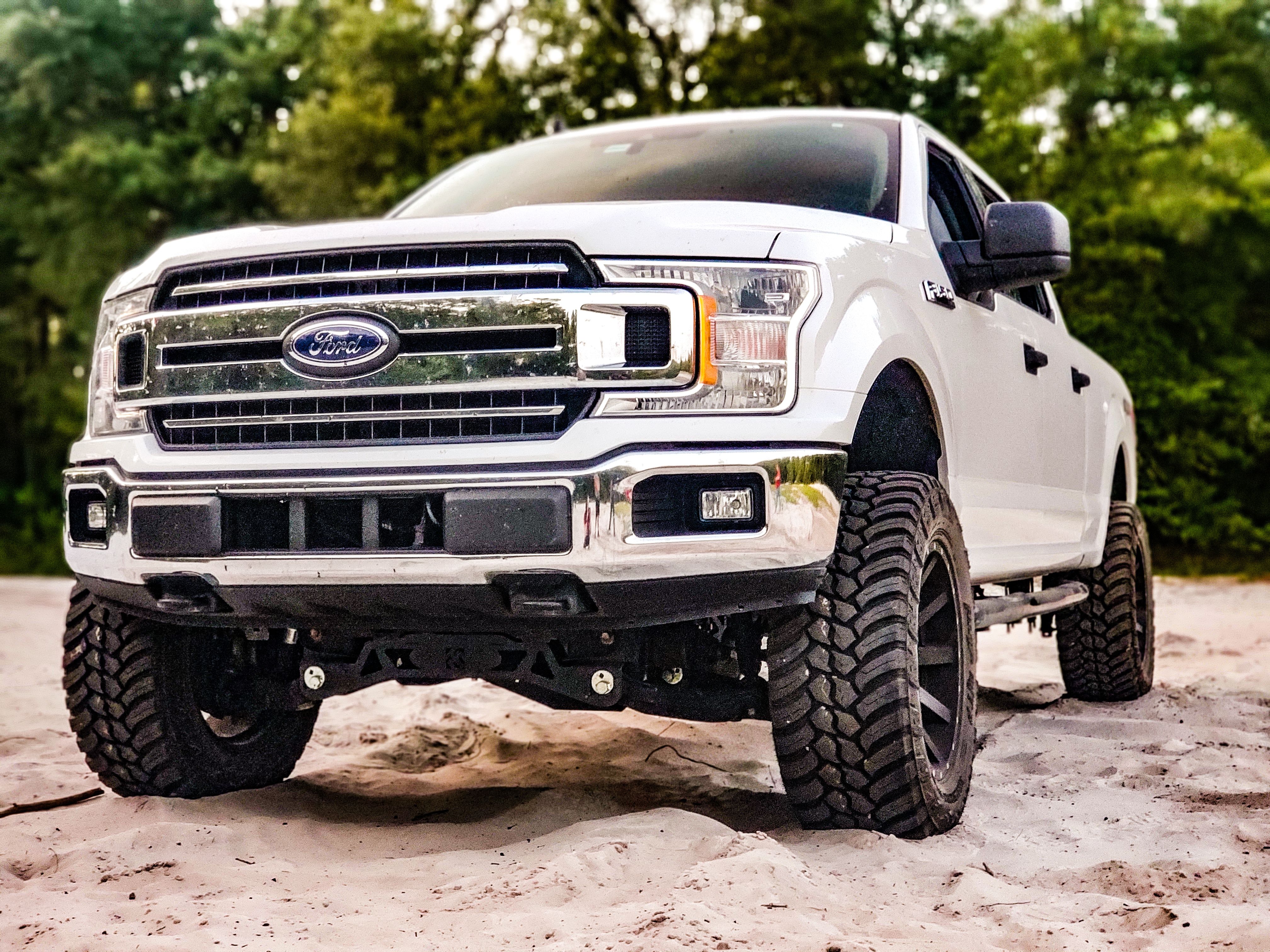White truck with bold grille parked on sandy surface, surrounded by lush trees.
