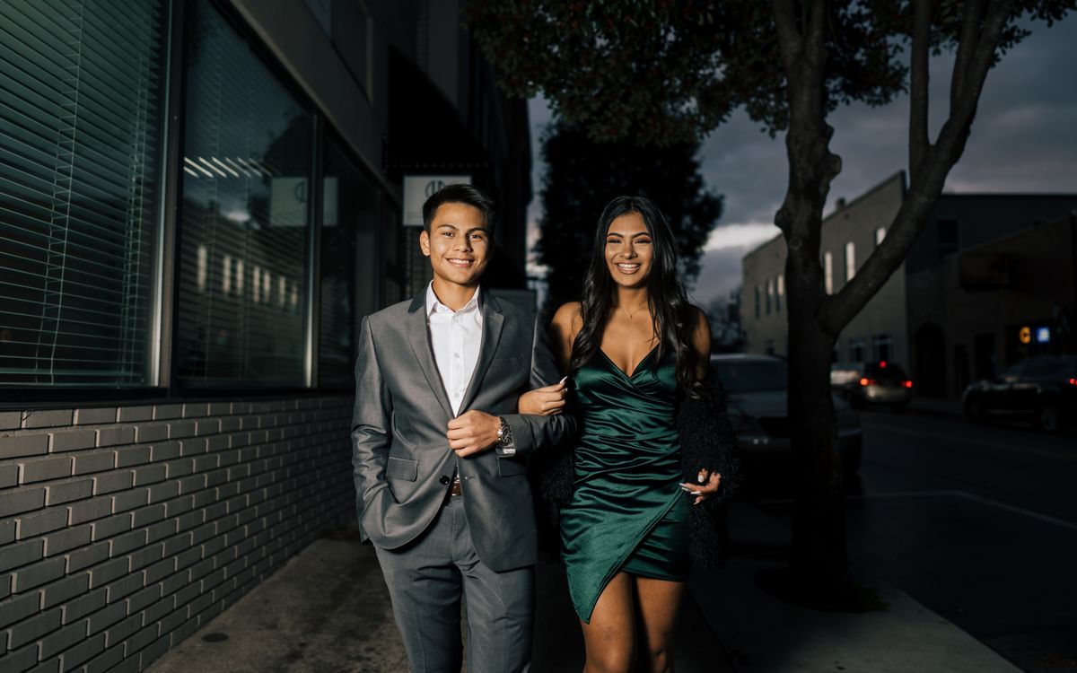 A stylish couple walking arm in arm on a city sidewalk at night, dressed in formal attire.