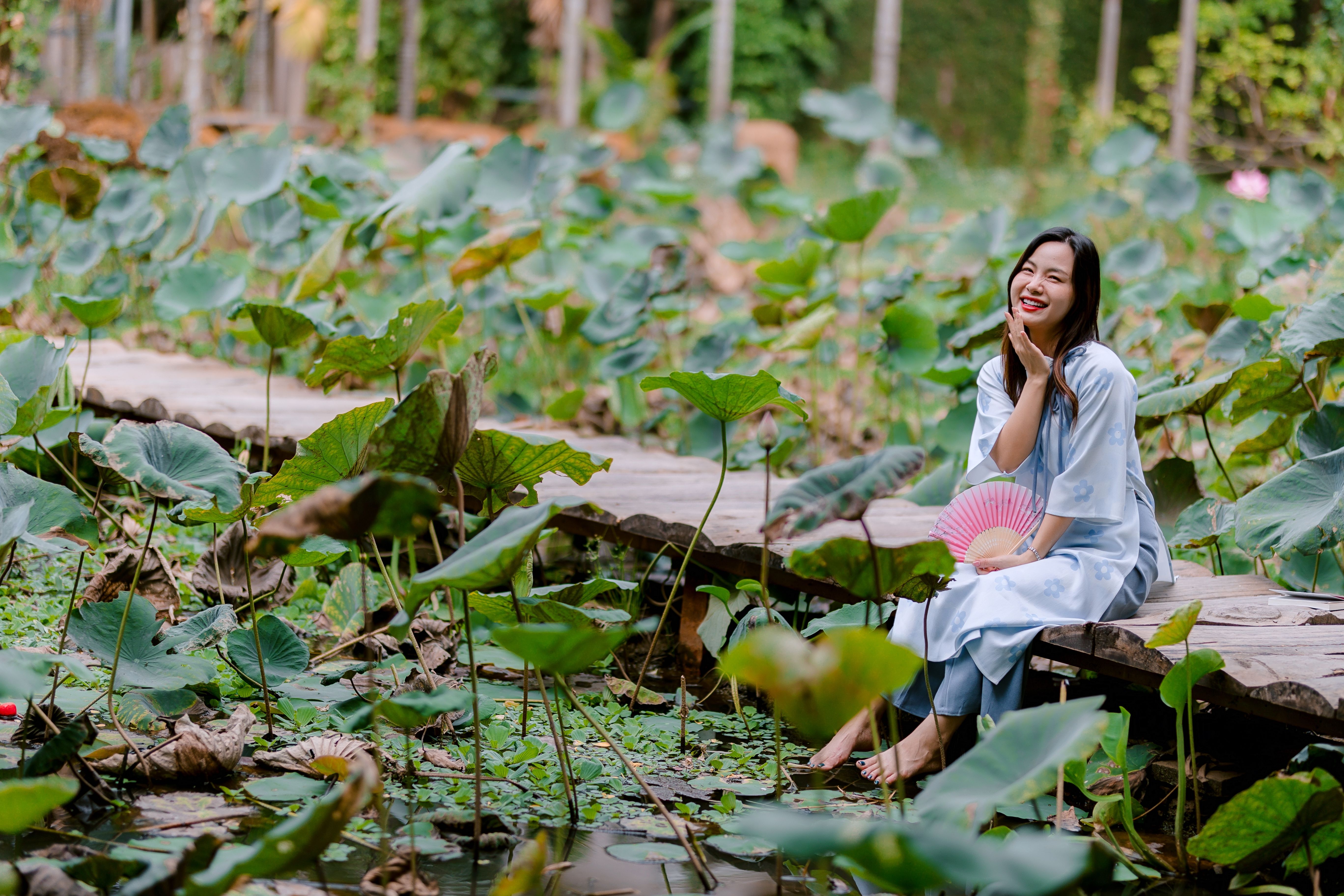 A young woman enjoys a serene moment by a lotus pond, holding a fan amidst lush green foliage.