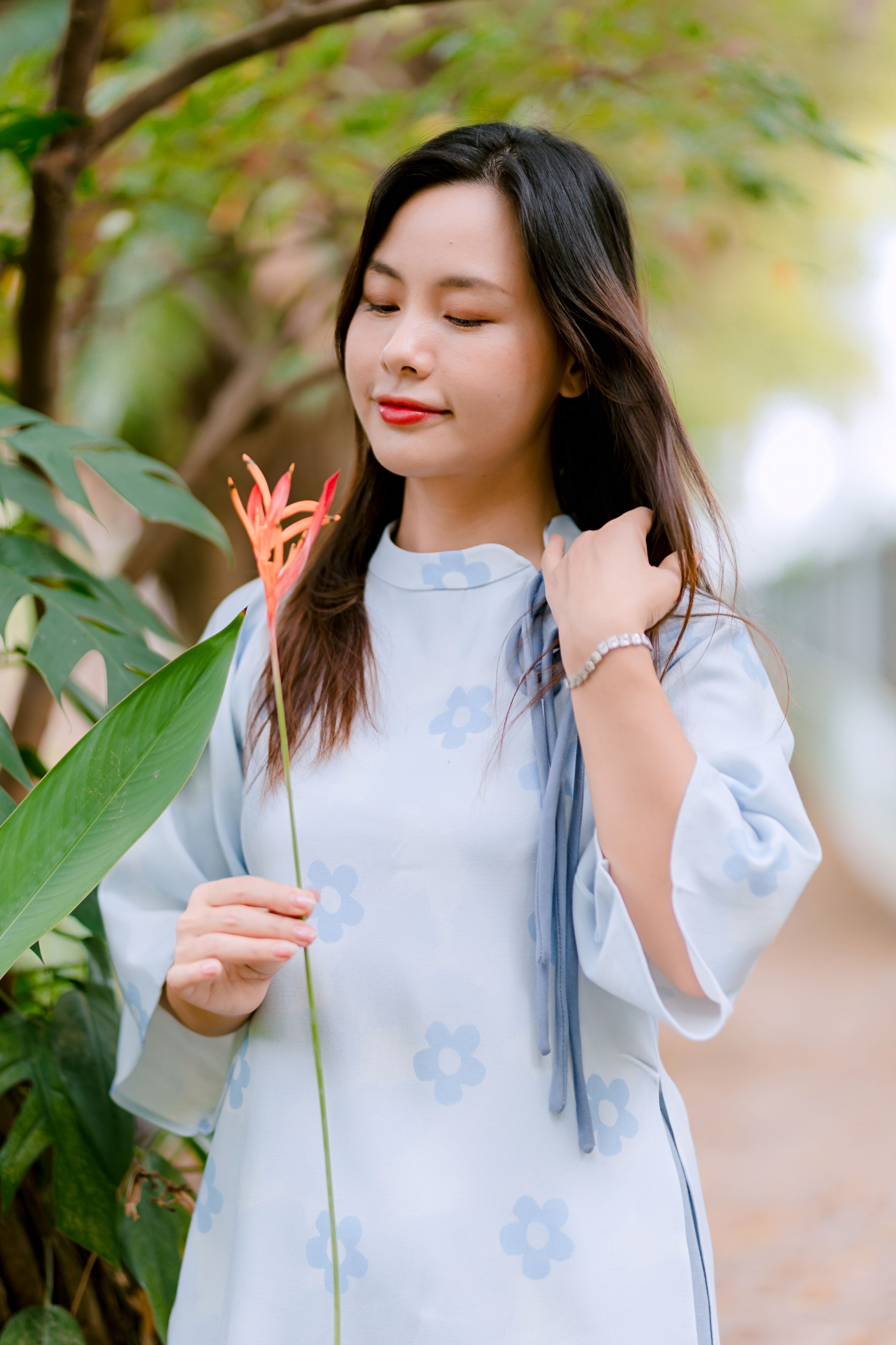 A woman in a light blue floral dress holds a bird of paradise flower outdoors.