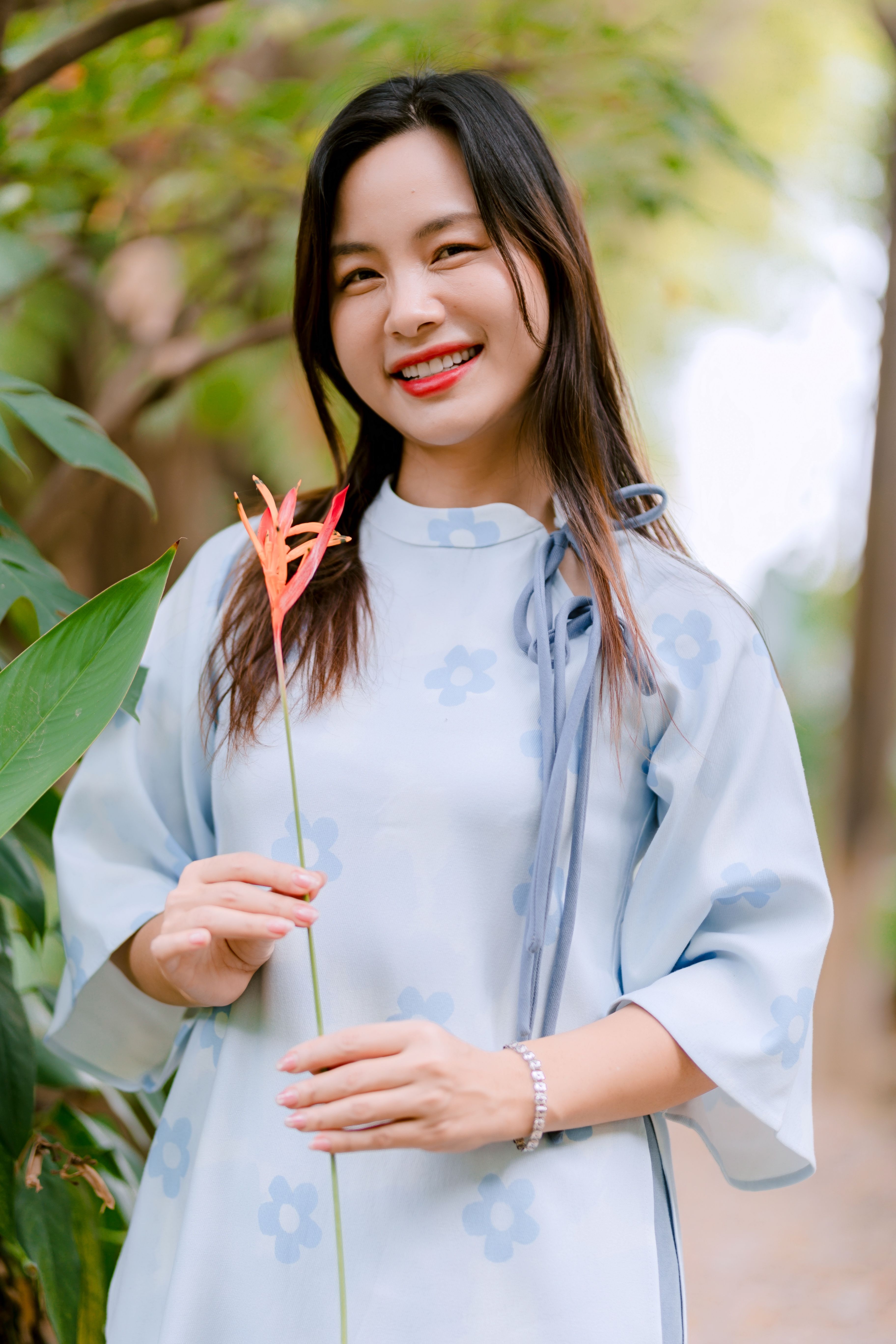 A cheerful woman in a light blue dress holding a vibrant flower outdoors.