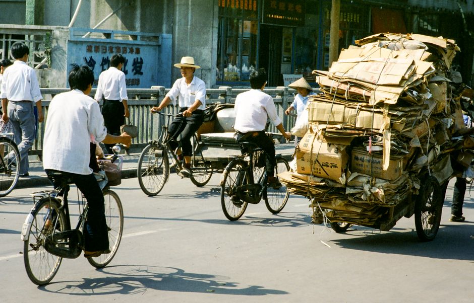 Cyclists navigate urban street with loaded cart in busy city scene.