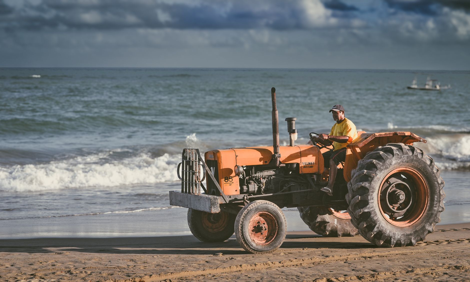 A man driving a tractor along the sandy shores of Recife, Brazil with waves in the background.