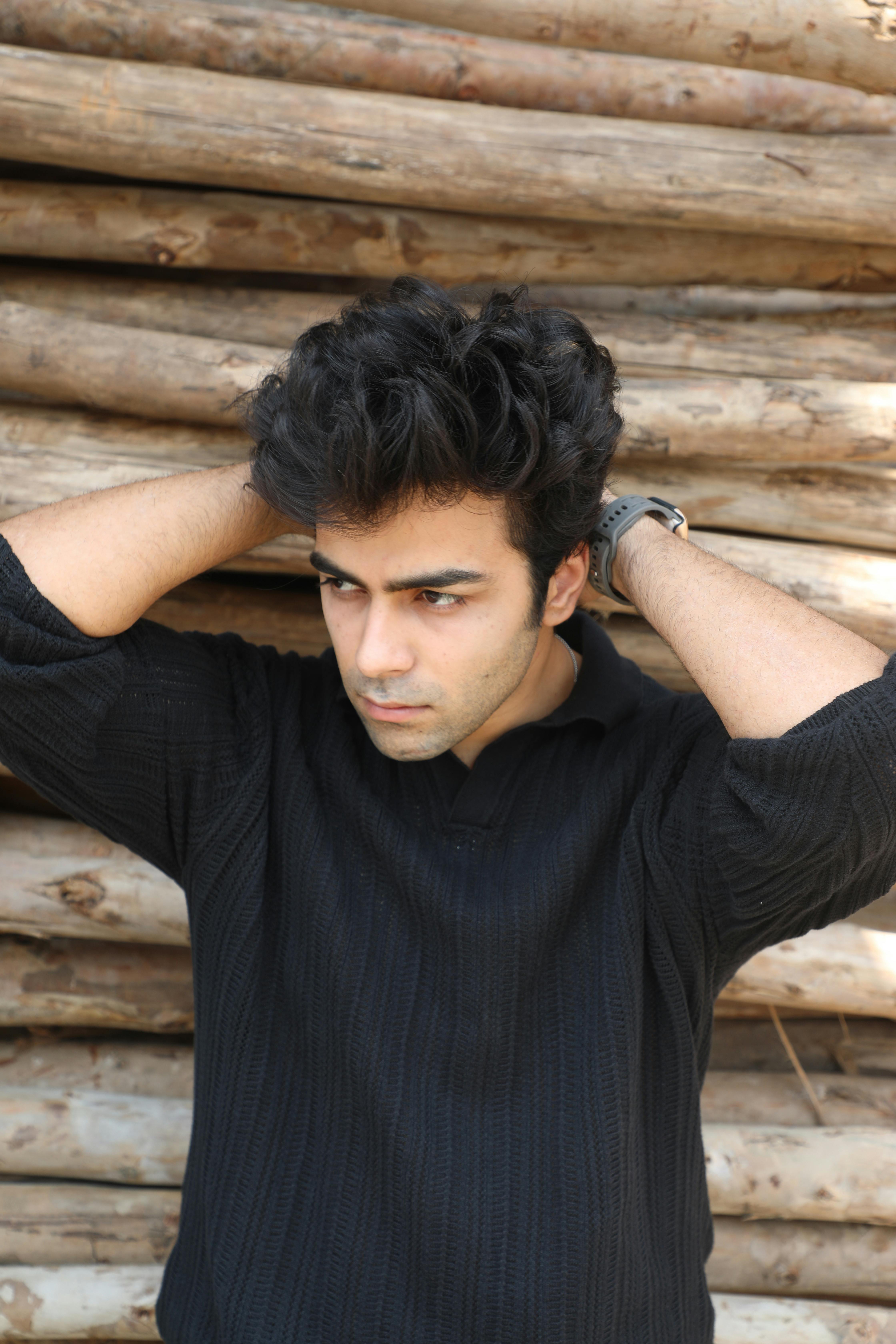 Stylish young man in black sweater, hands on head, in front of stacked logs outdoors.