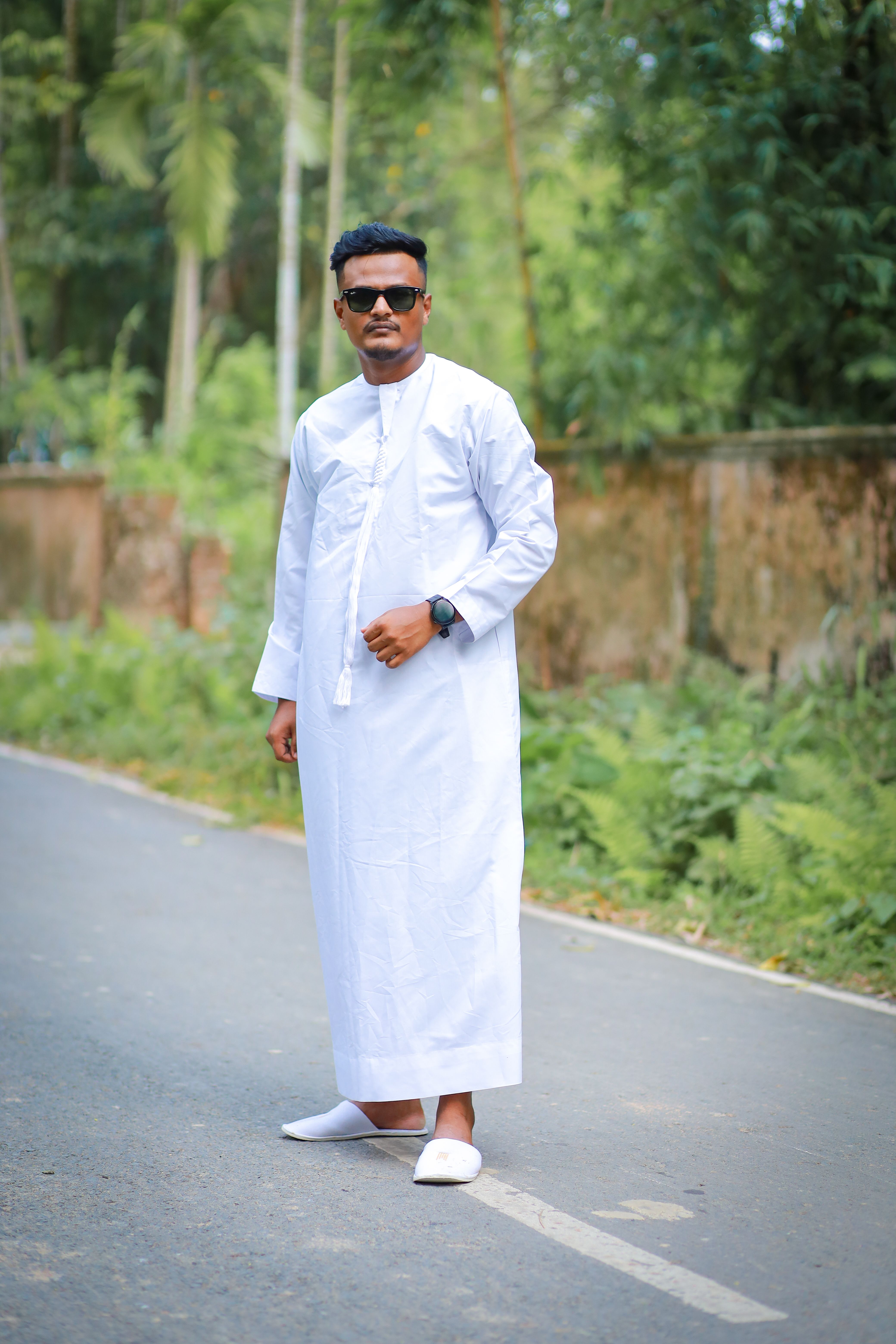 Confident man in white thobe posing on a forest road, showcasing traditional attire.
