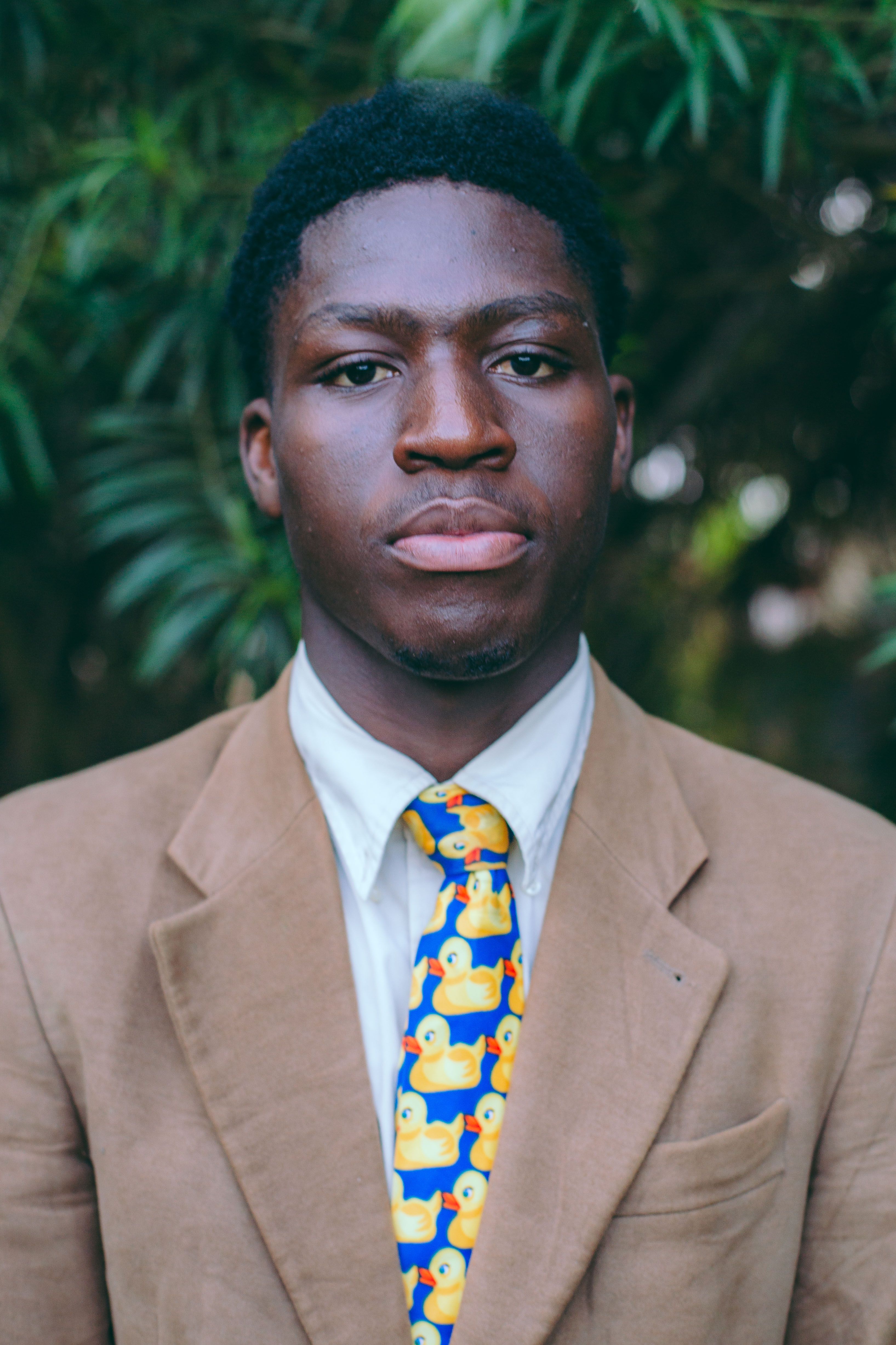 Close-up portrait of a young man wearing a brown suit and a colorful duck-patterned tie, outdoors.
