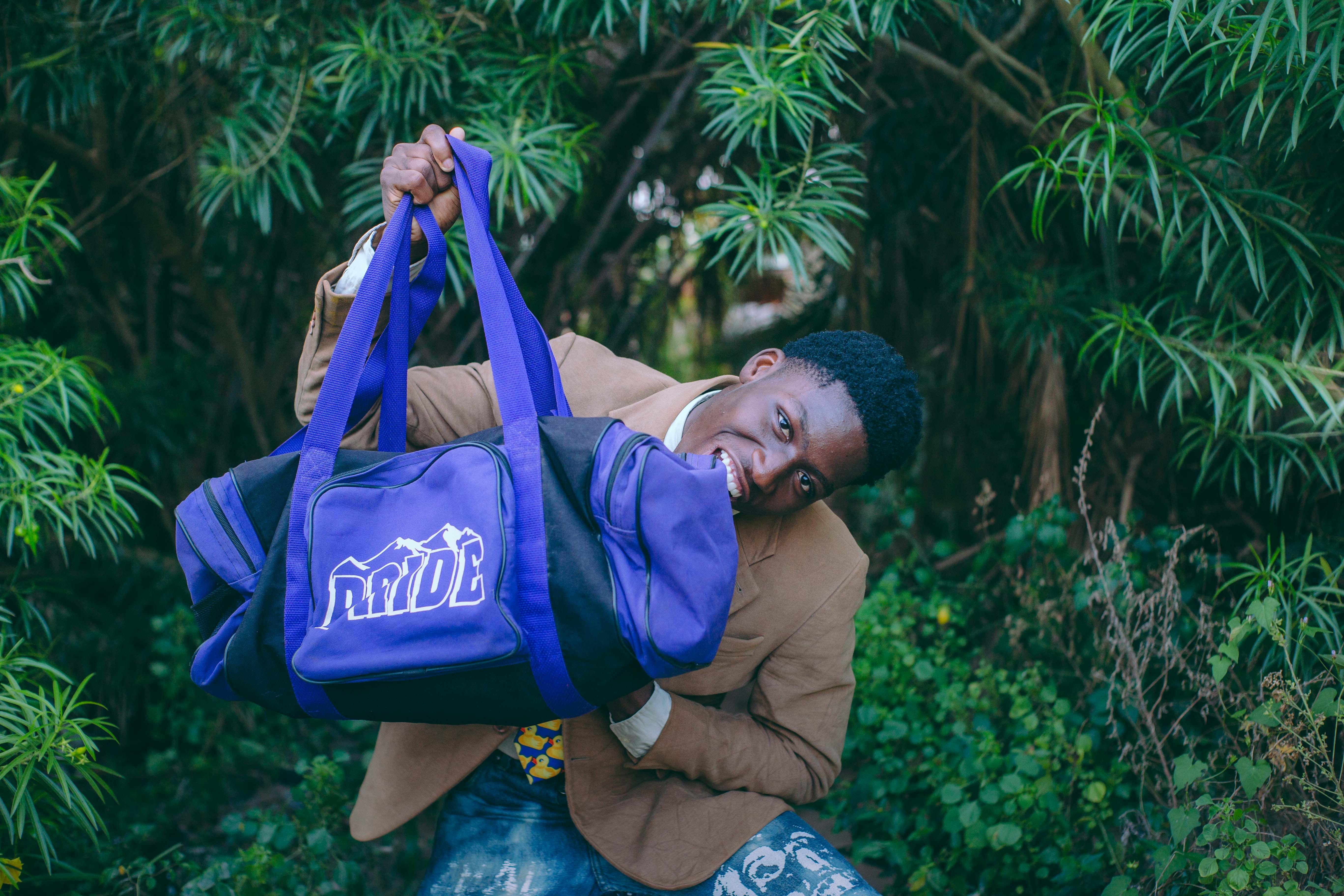 Young man playfully holding a purple duffel bag in a lush, green outdoor setting.