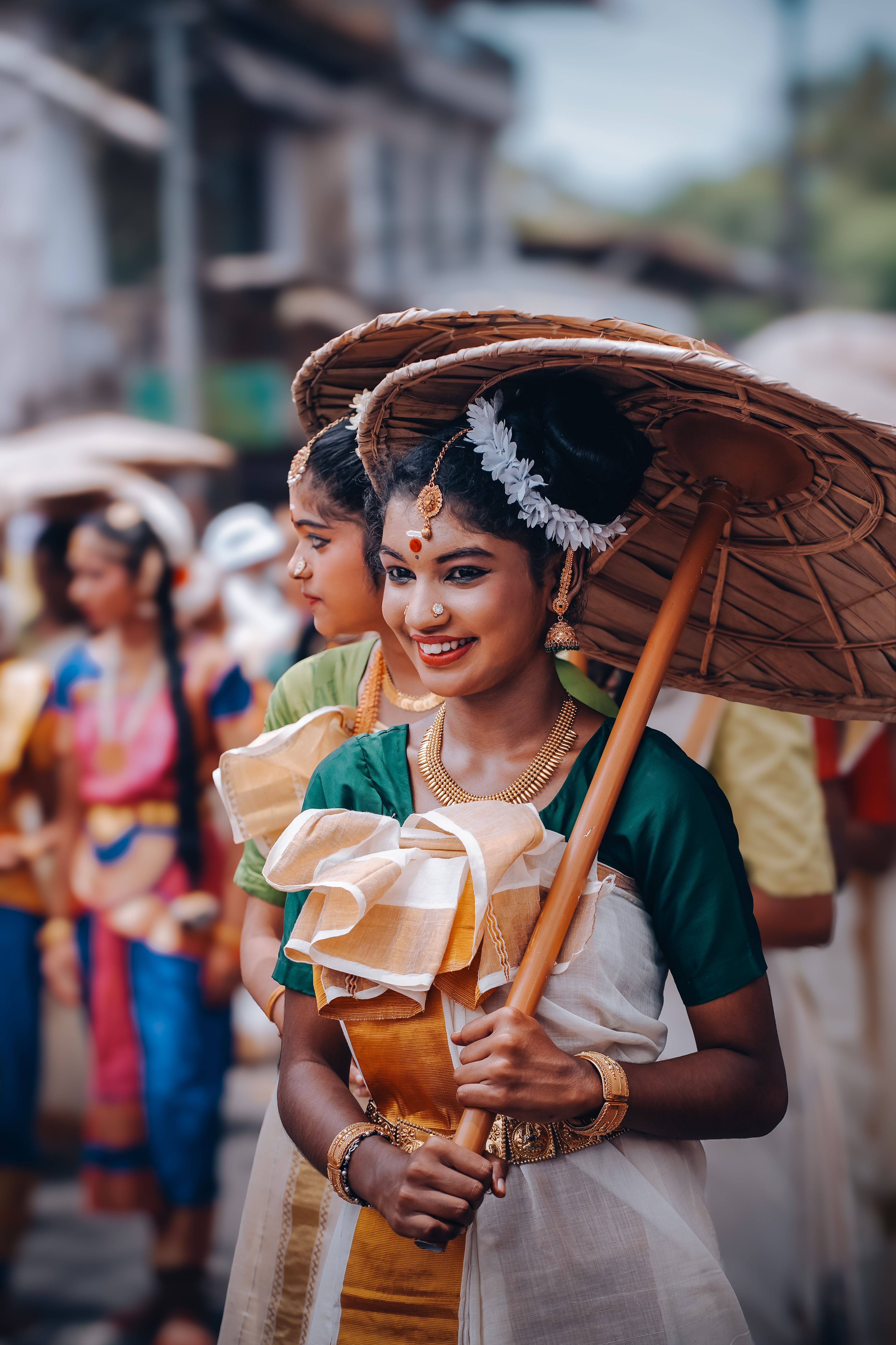 Young women in vibrant traditional attire with umbrellas during a cultural parade in KL, India.