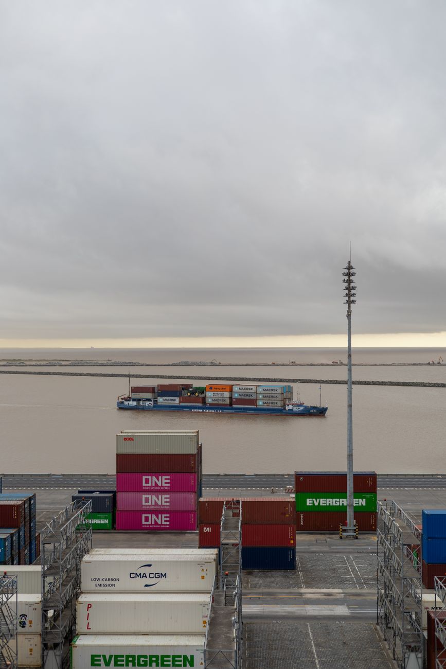 Een vrachtschip vaart door een drukke haven, omringd door kleurrijke zeecontainers.