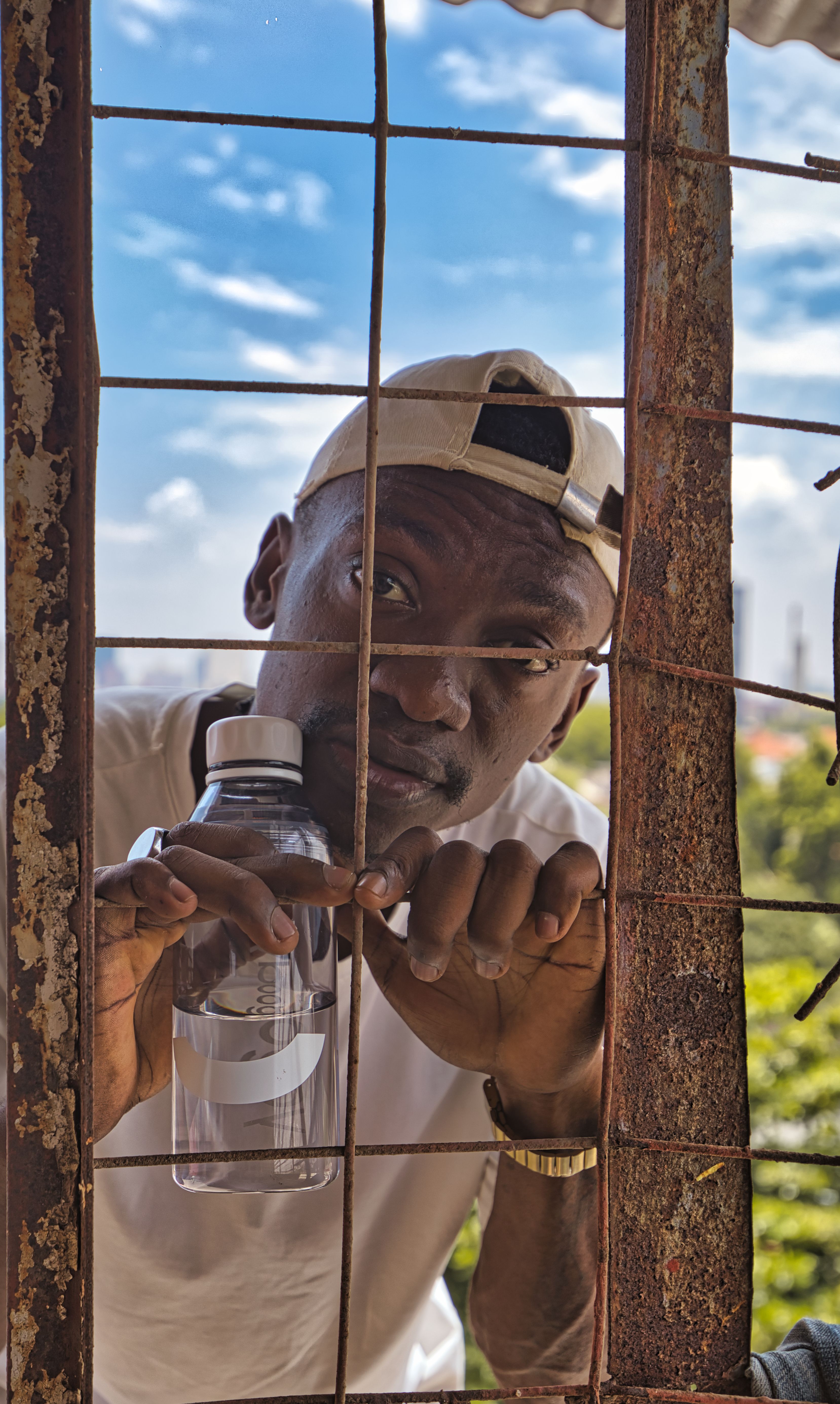 A man peeks through a rusty fence holding a water bottle on a sunny day in Luanda, Angola.