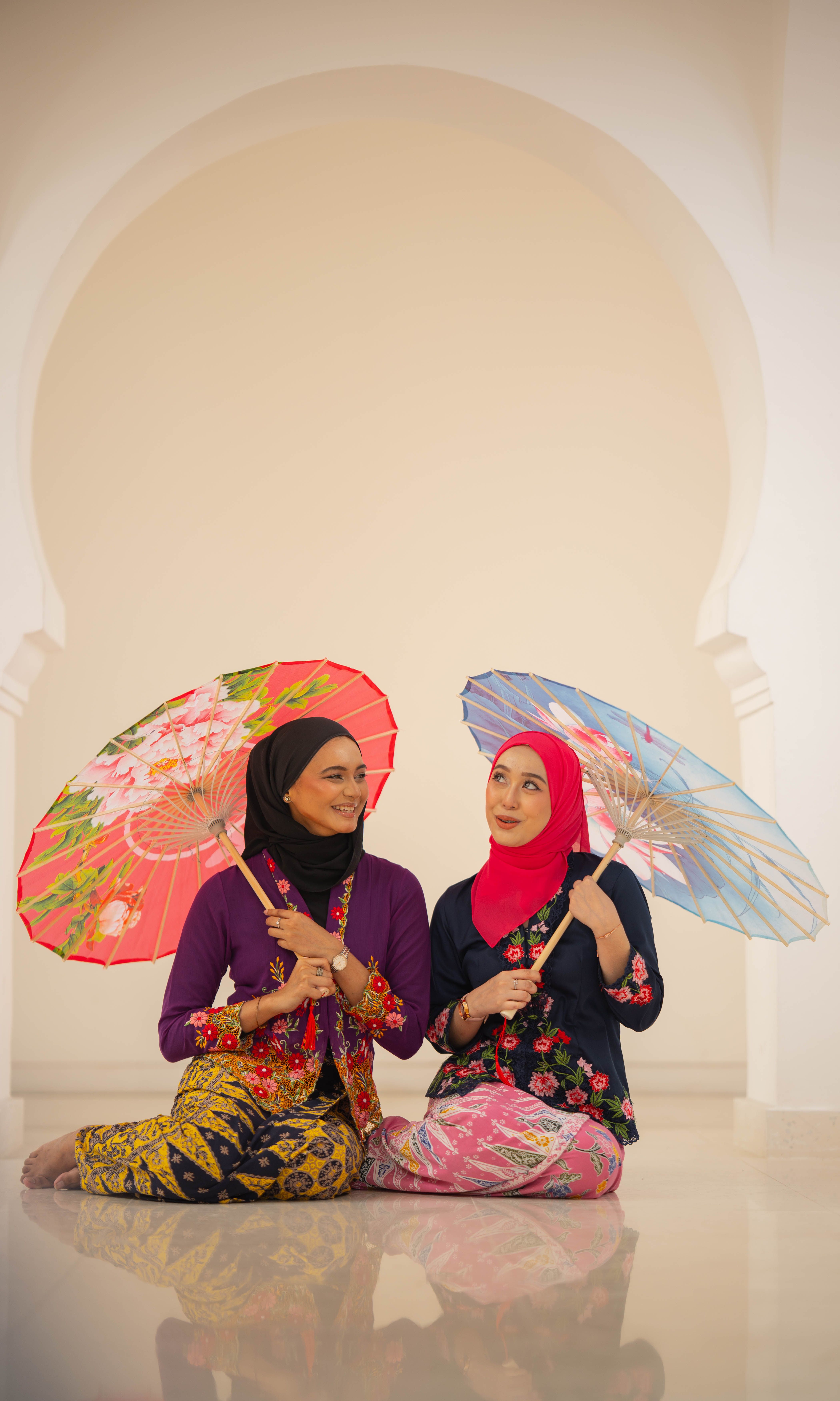 Two women in traditional attire sit indoors with colorful parasols.