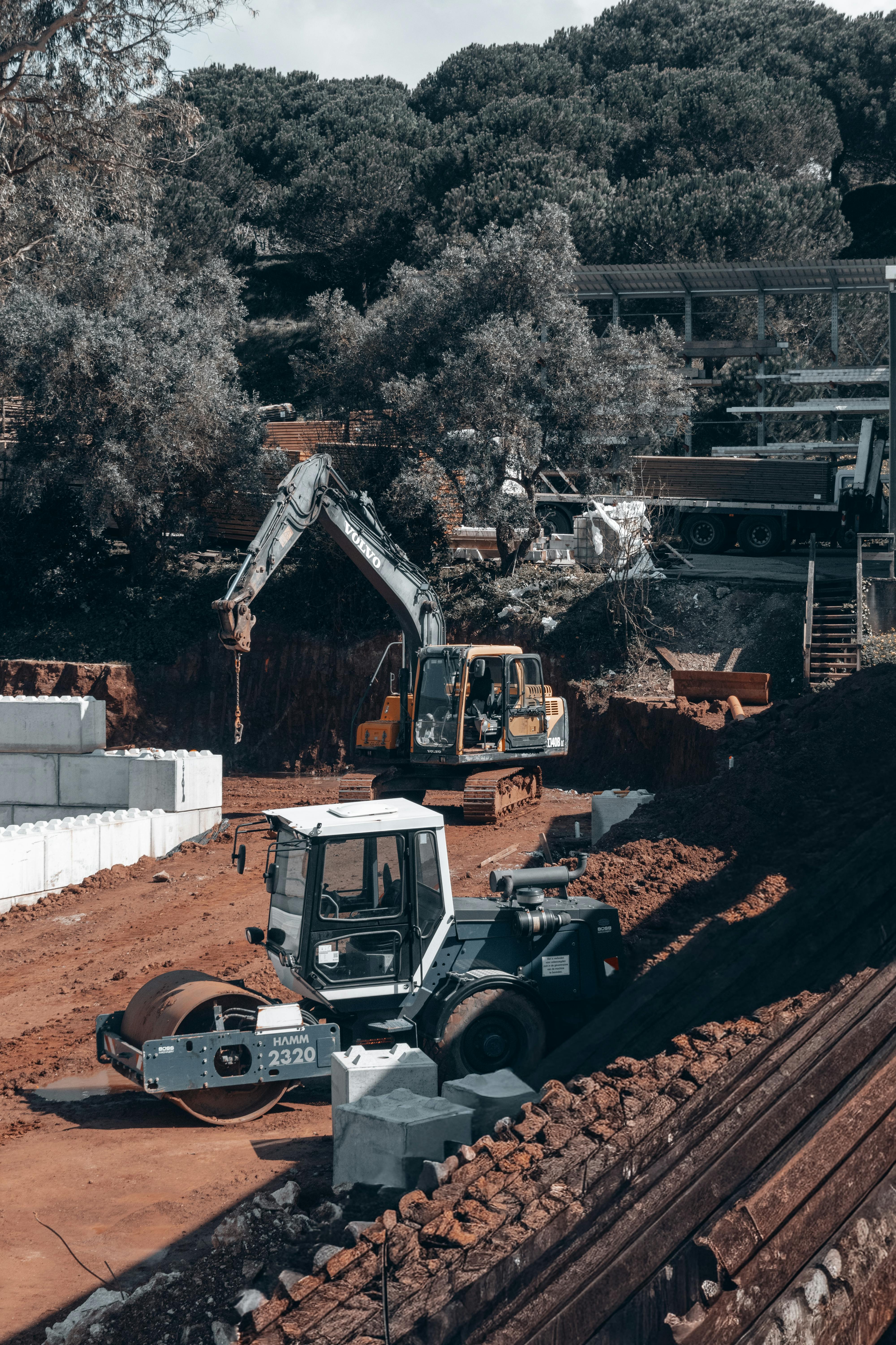 Excavators and vehicles operate on a forested construction site under a clear sky.