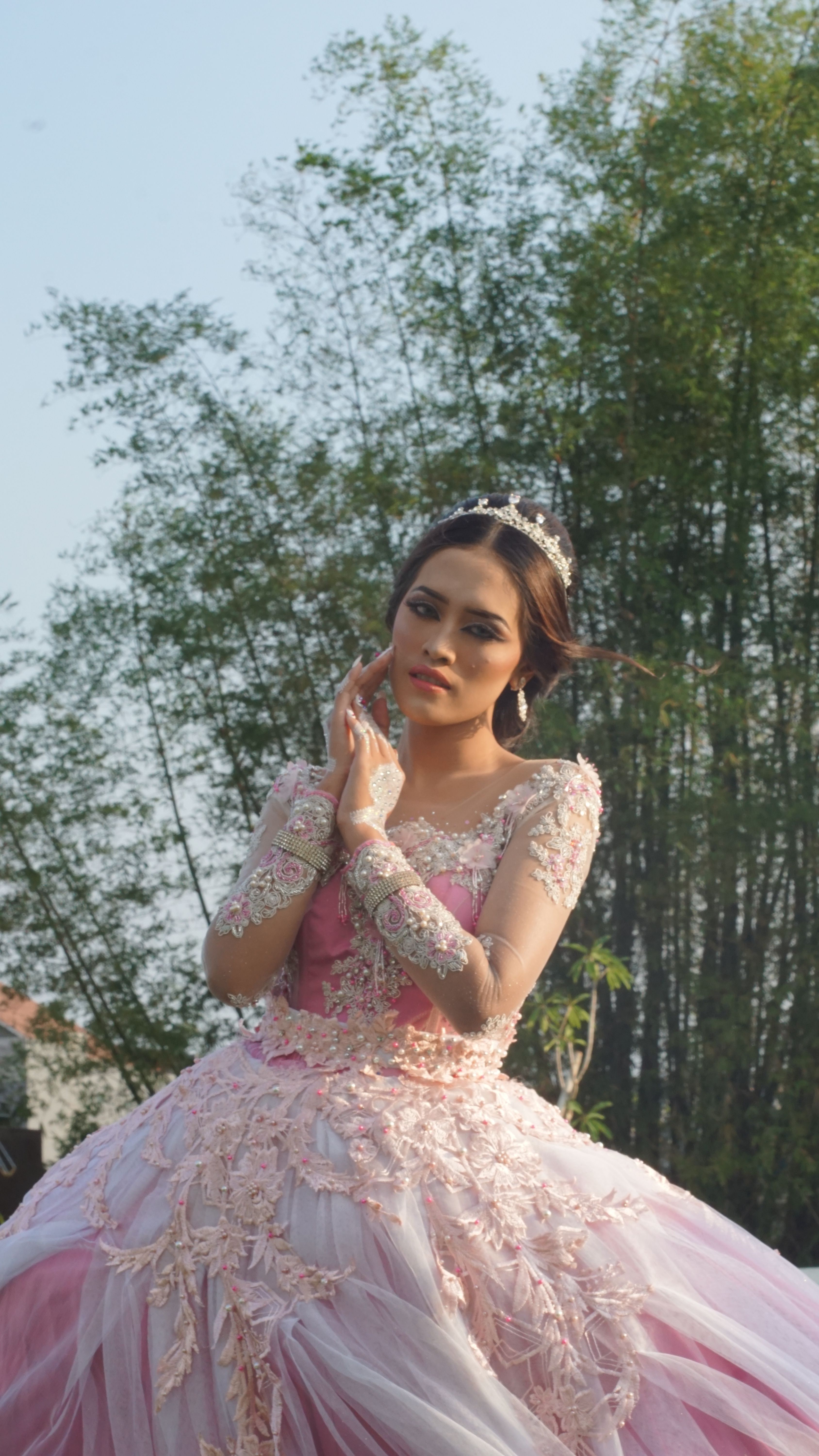 Portrait of a woman in an elegant, pink gown with a tiara, set outdoors against a bamboo backdrop.