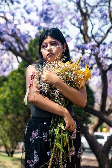 Fotomural Un niño en un vestido floral sostiene un ramo de flores bajo árboles de jacarandá.