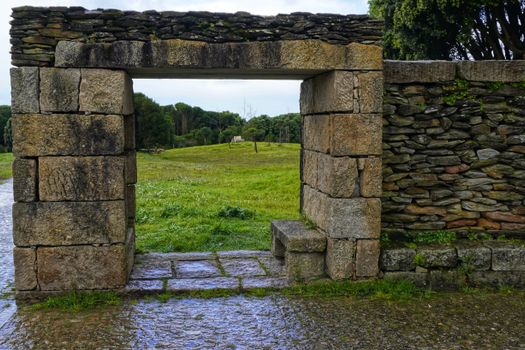 A rustic stone gateway opens to a lush green meadow surrounded by trees.