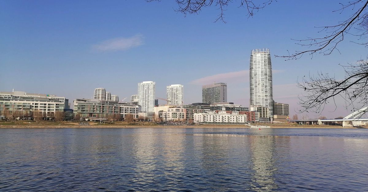 View of modern city skyline with buildings reflecting on river under clear blue sky.