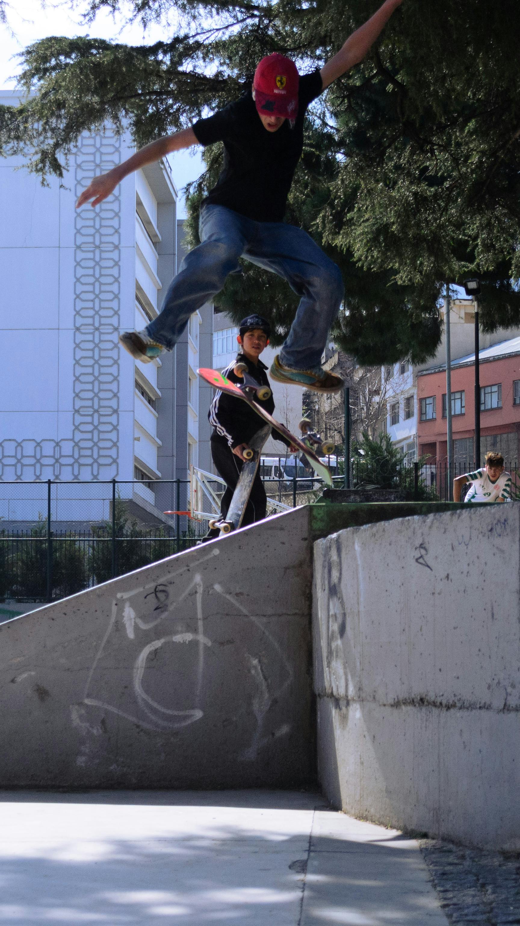 A teenager performs a skateboarding jump in an urban skate park setting amidst city buildings.