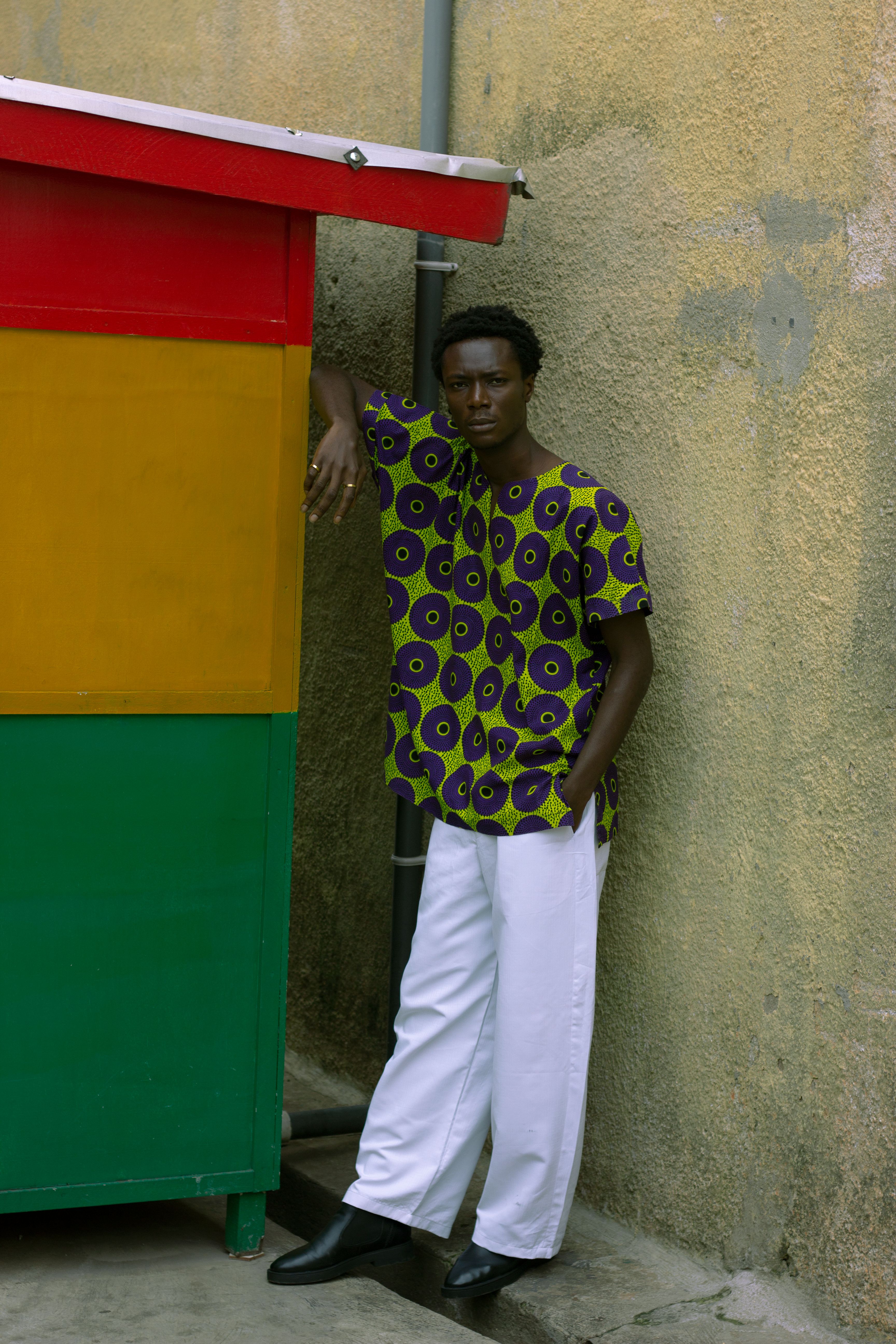Stylish man in vibrant African print shirt and white pants leaning against a wall.