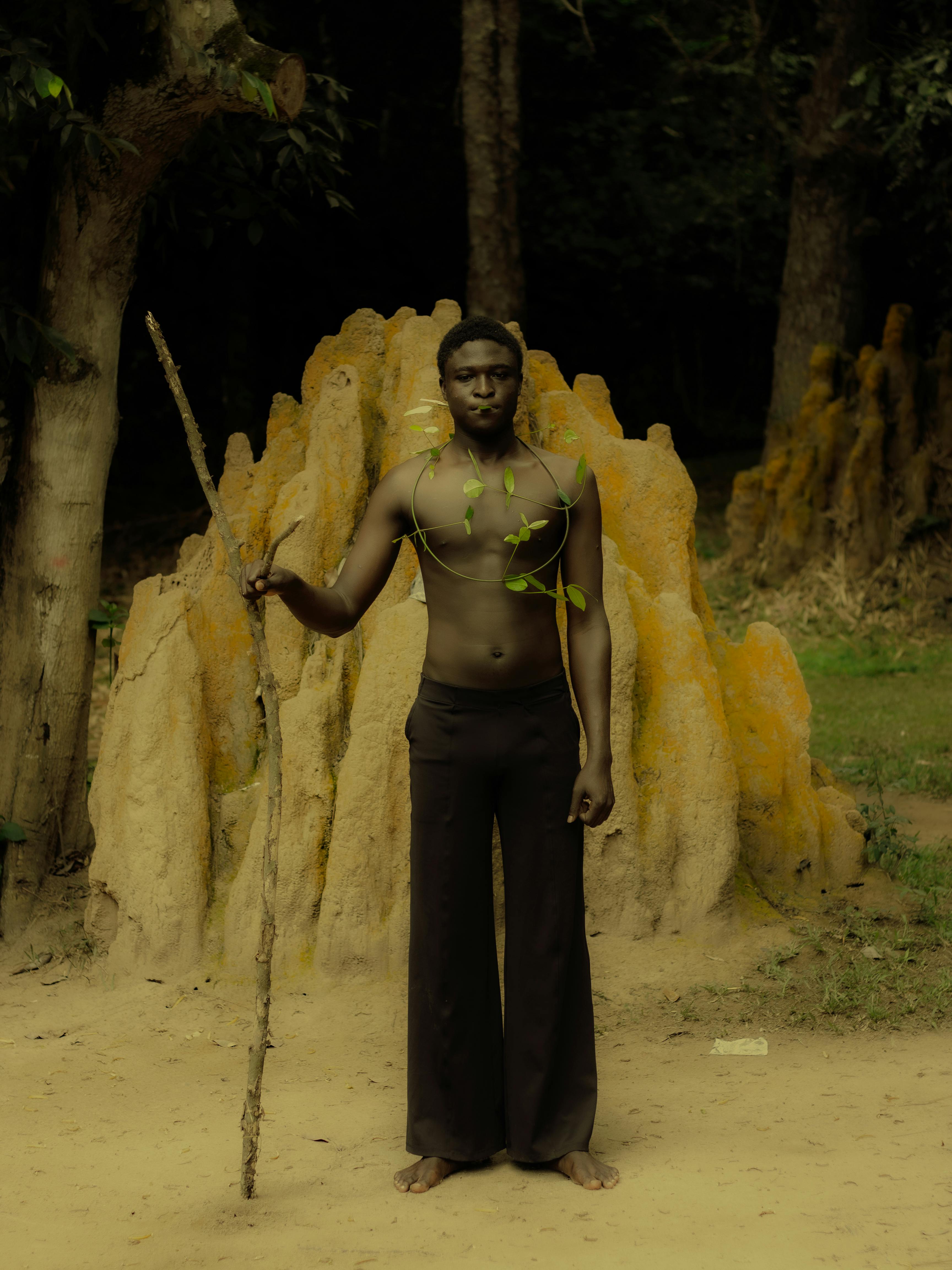 Man in nature holds stick, stands by termite mound. Natural elements surround.