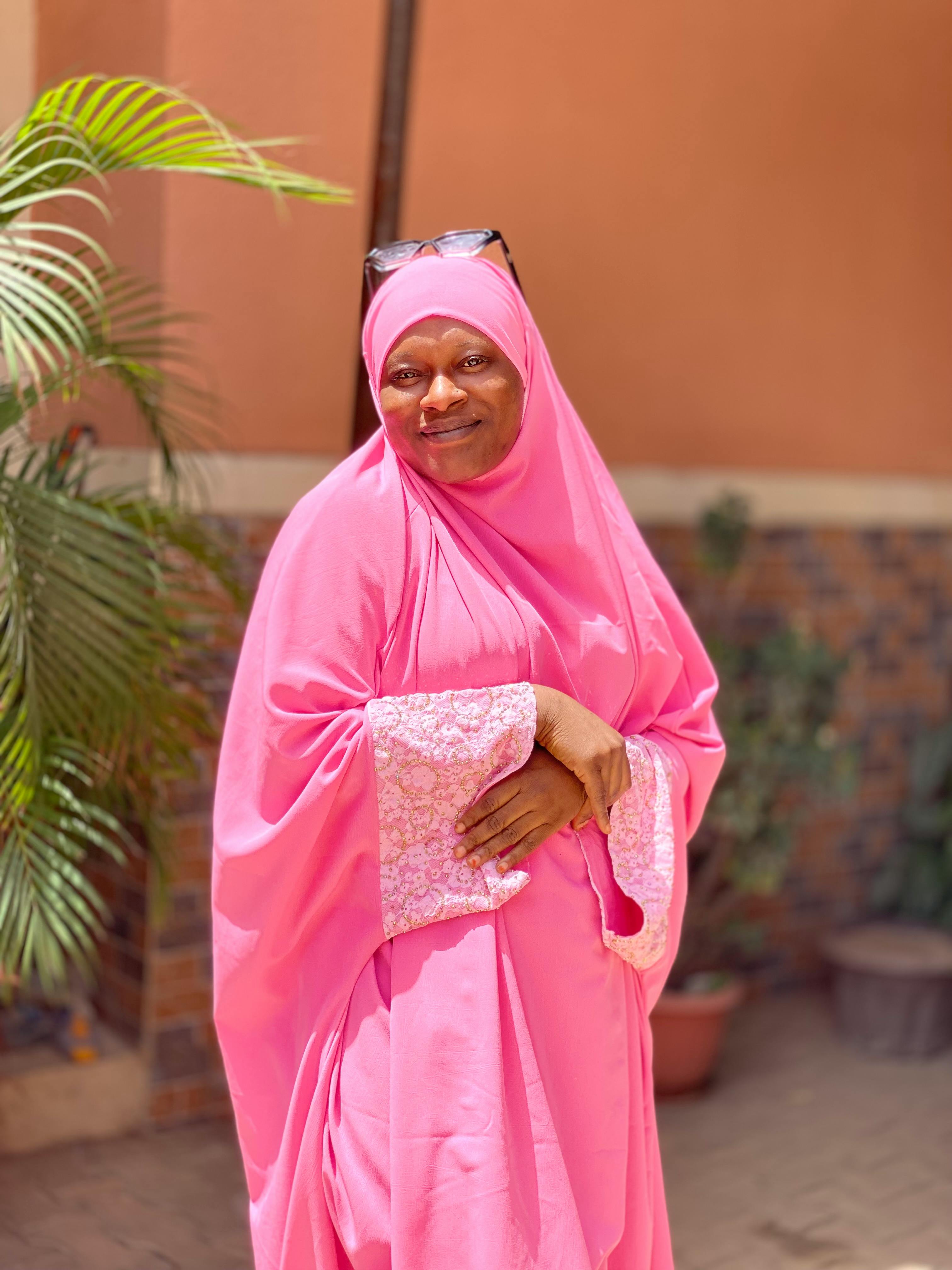 A joyful woman in a bright pink hijab stands outdoors, exuding warmth and confidence.