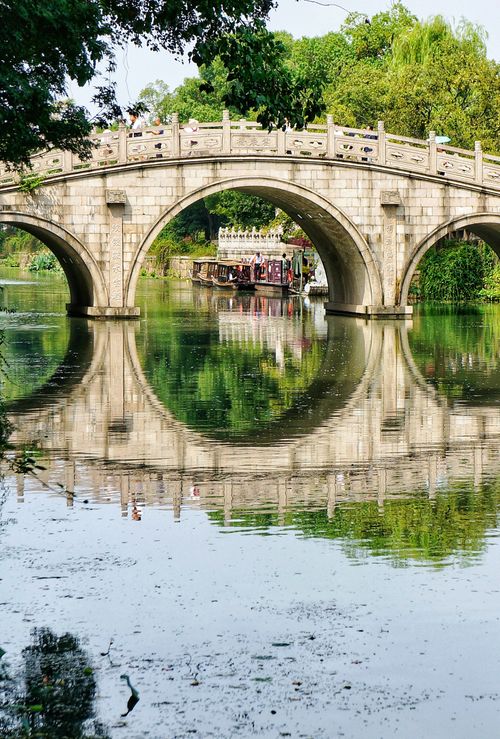 Free A picturesque stone arched bridge reflecting over a tranquil river surrounded by lush greenery. Stock Photo