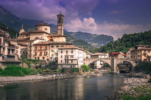Free Charming Italian village with classic architecture, scenic river, and stunning mountain landscape. Stock Photo