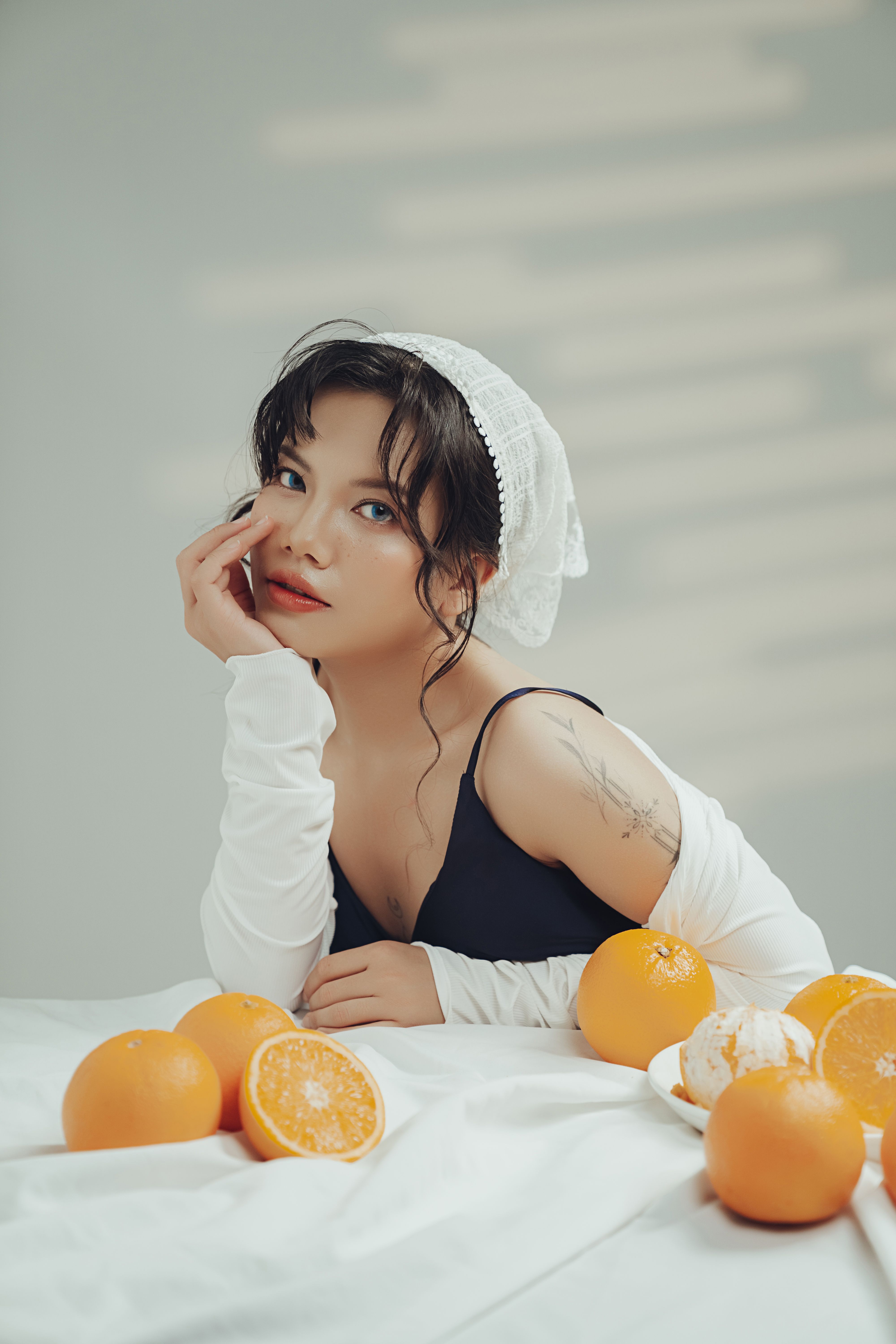 A stylish woman with oranges on a table, bathed in soft lighting indoors, creating a calm and elegant atmosphere.