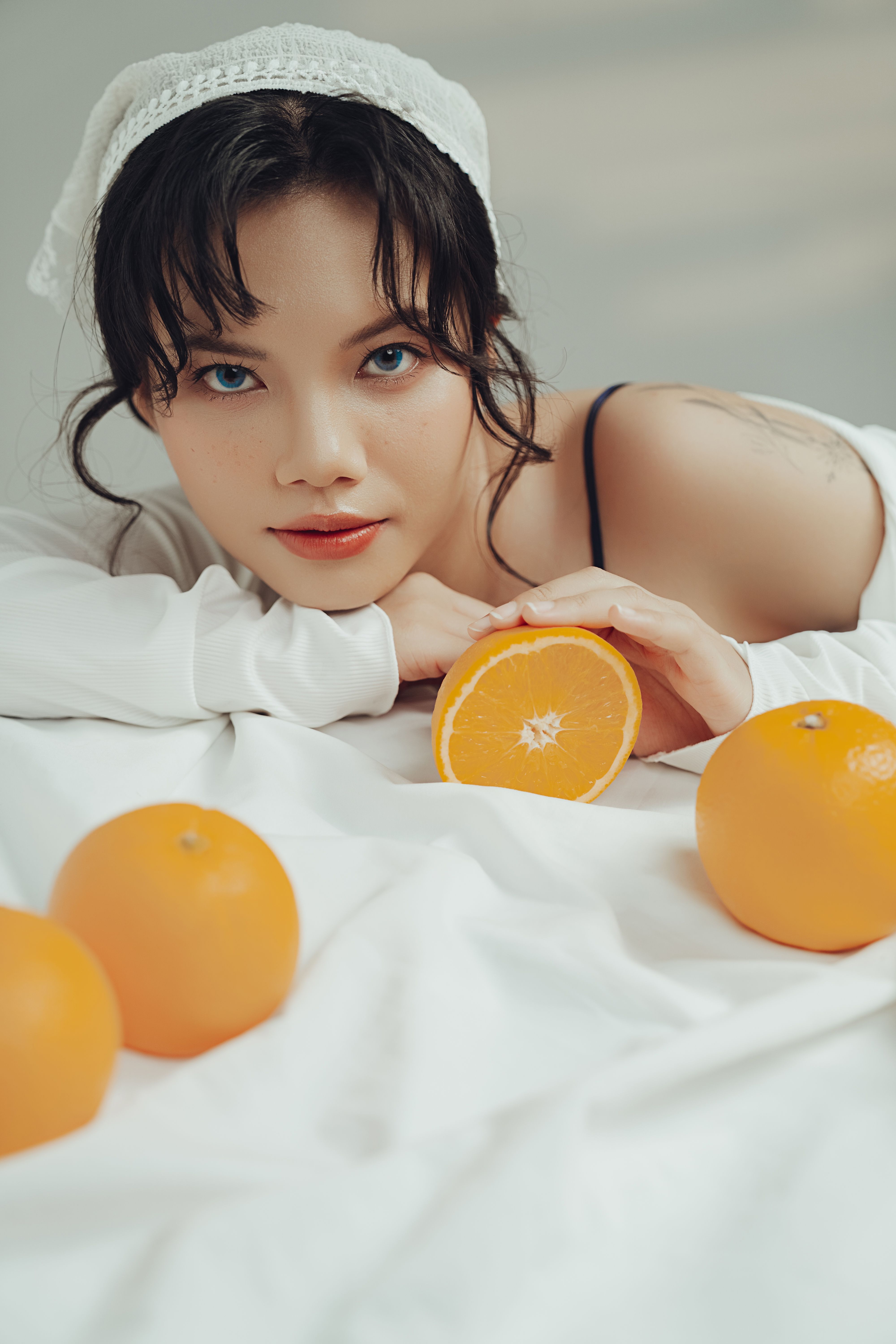 A woman with dark hair poses with oranges, lying on a white sheet, creating a fresh and serene look.
