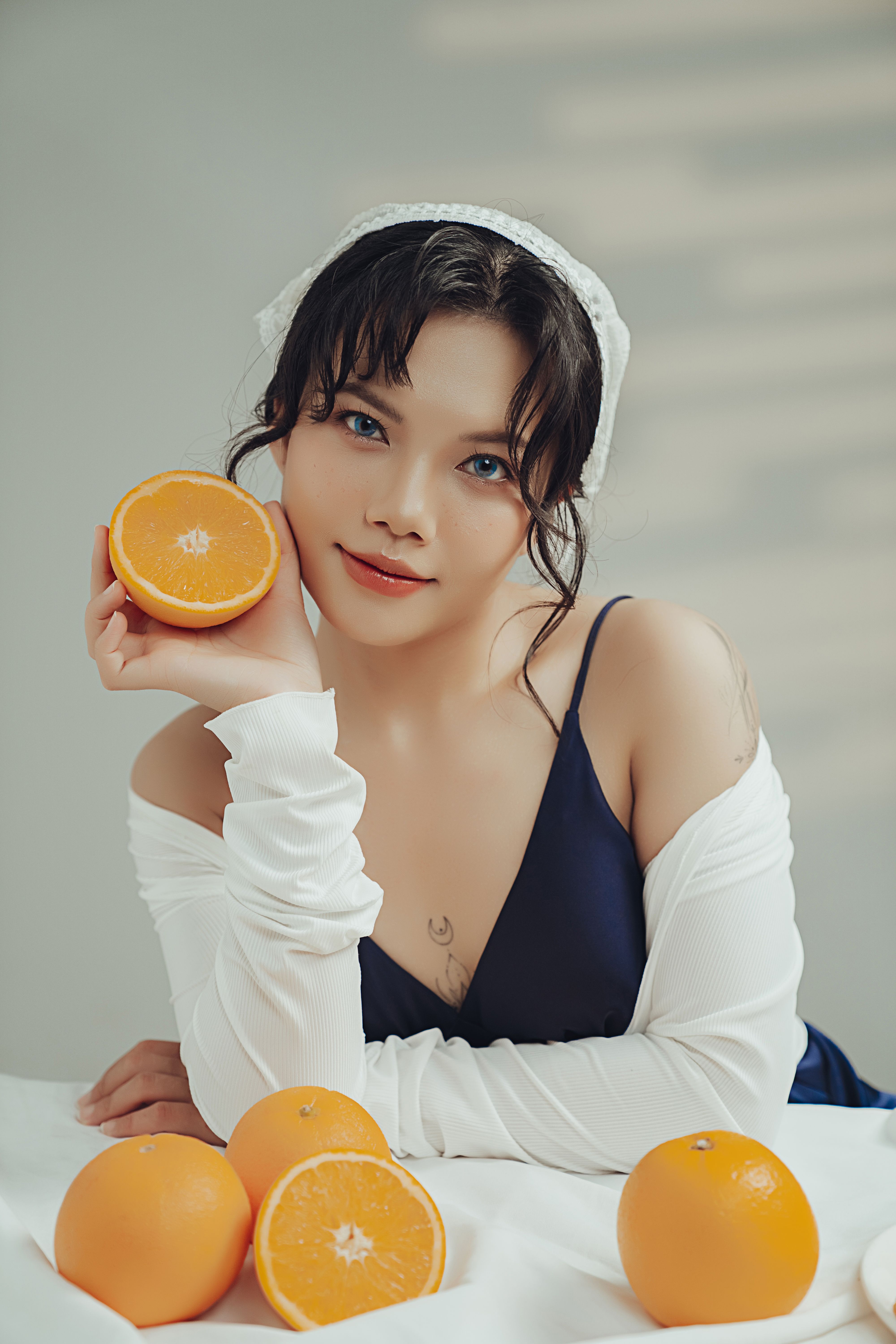 Young woman in white headscarf holding a sliced orange while surrounded by whole oranges on a table.