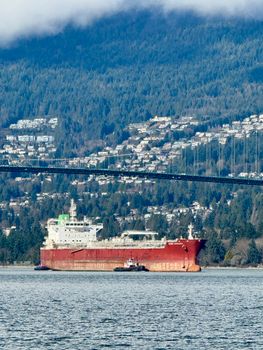 A cargo ship sails under a bridge in Vancouver, framed by lush forests and cityscape.