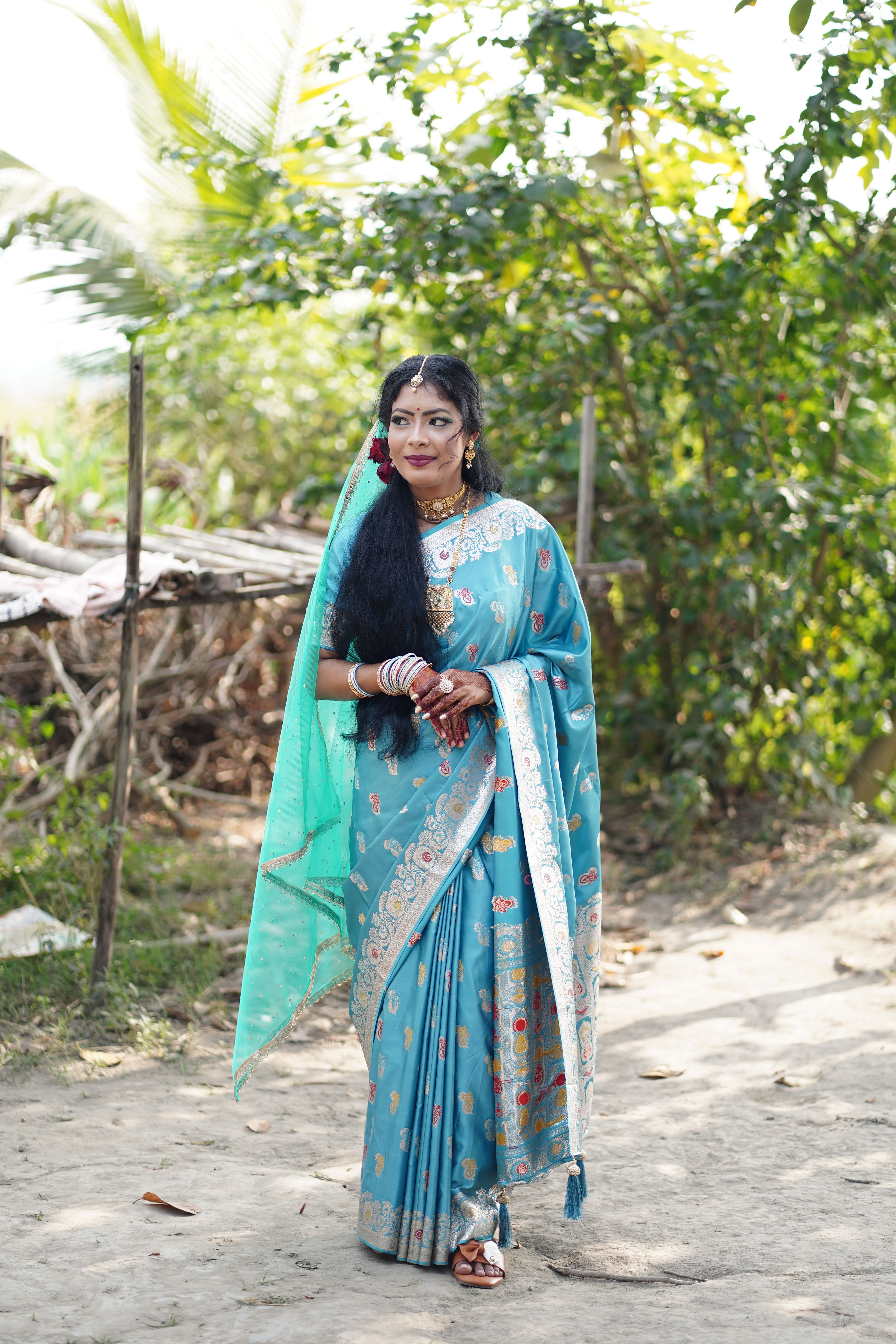 A woman wearing a vibrant blue sari stands gracefully in a natural outdoor setting.