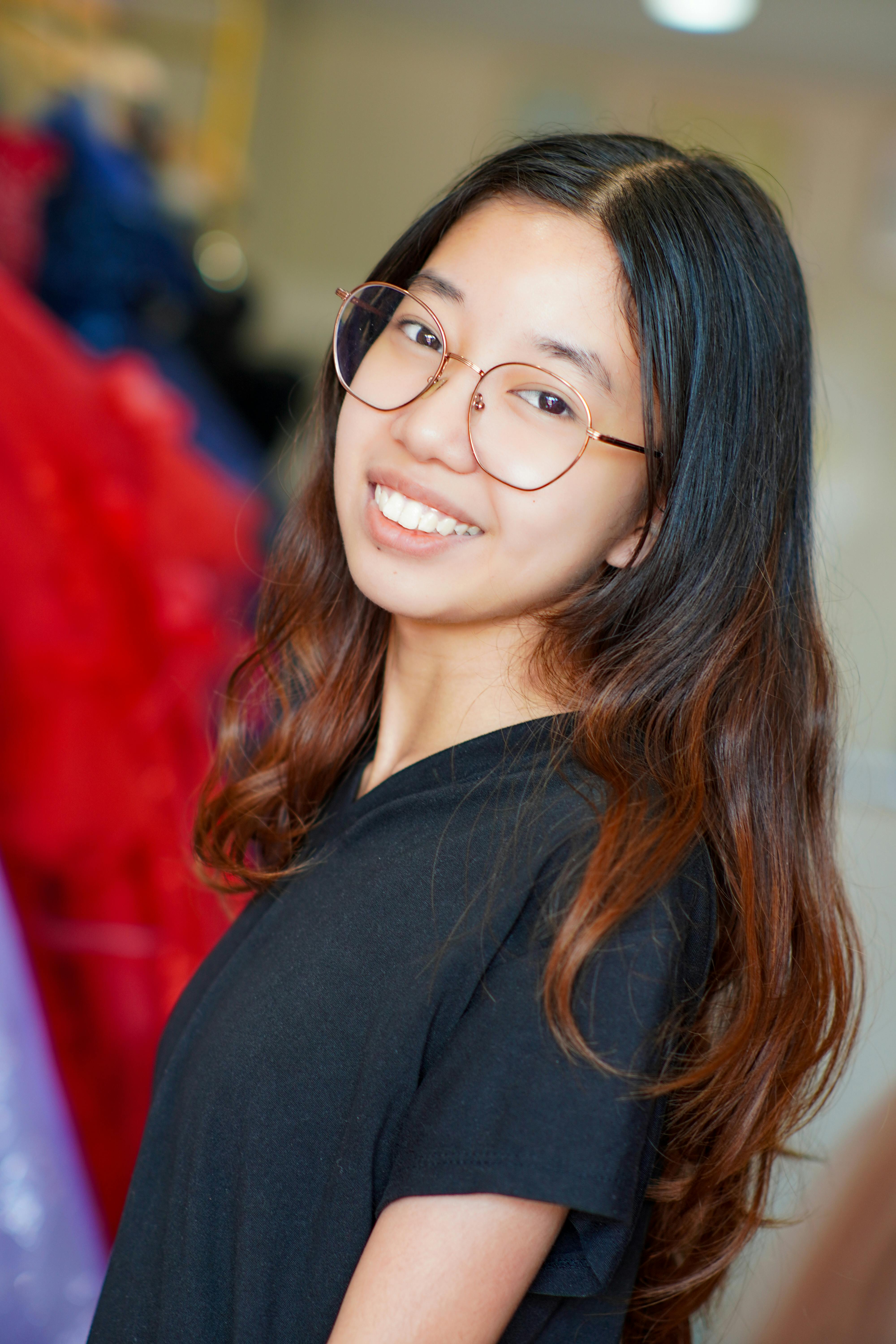 A cheerful young woman wearing glasses, smiling indoors with a blurred colorful background.