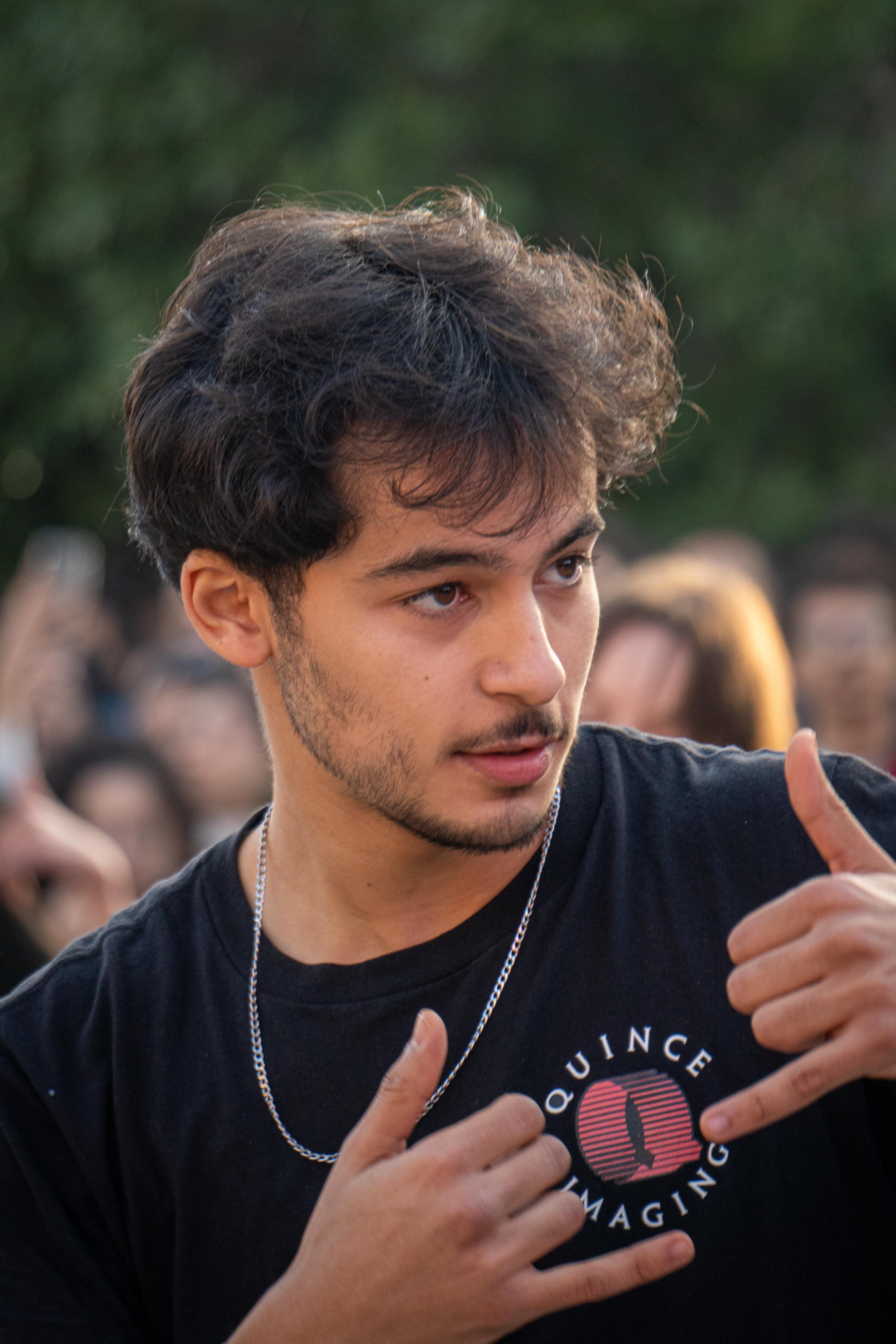 Portrait of a young man making gestures amidst a crowd outdoors, showcasing lively expression and vibrant atmosphere.