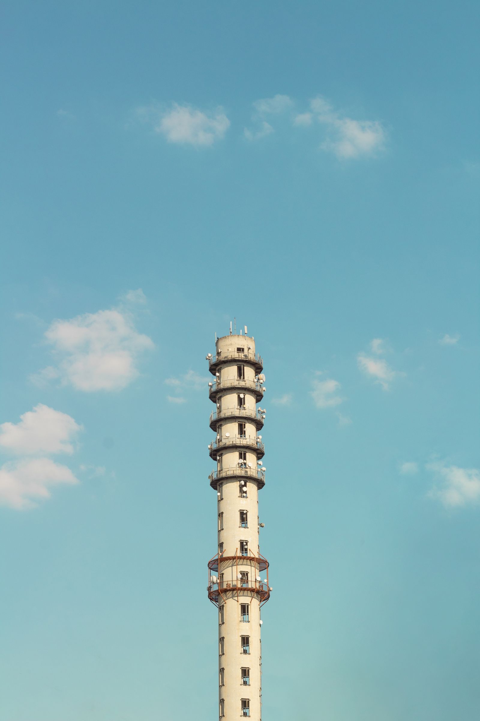 A tall white communication tower with stacked circular platforms and antennas rising into a clear blue sky with scattered clouds.