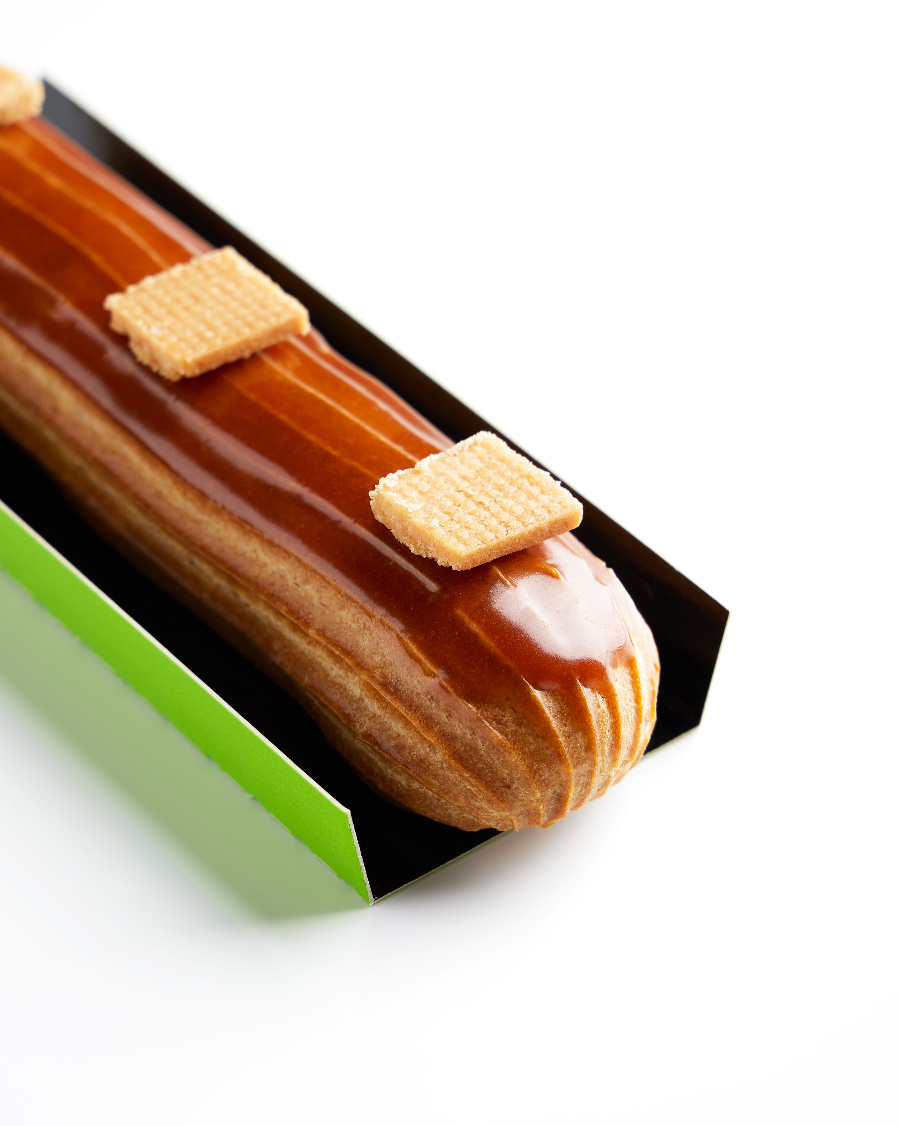 A beautifully glazed eclair topped with wafers in a green box on a white background.