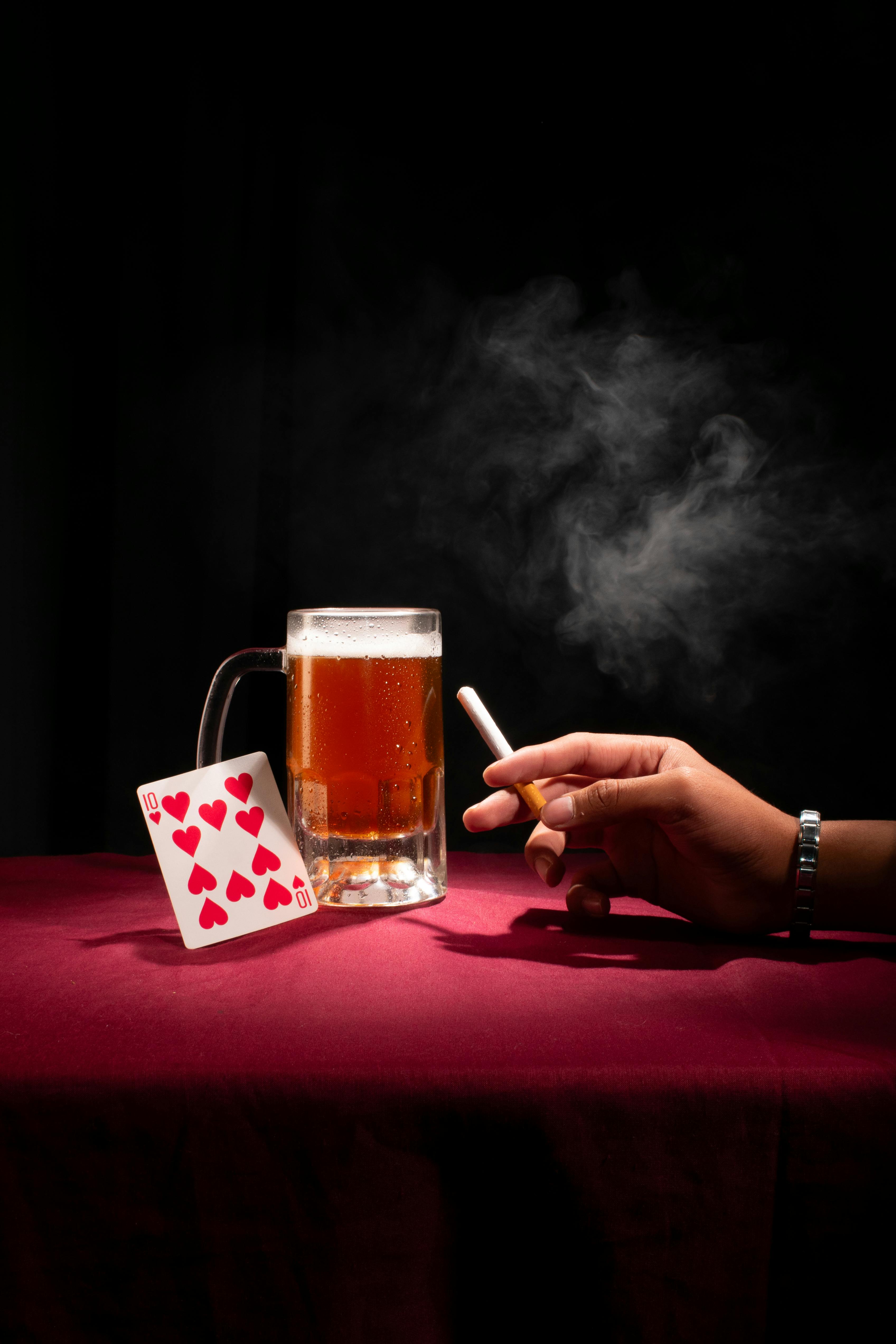 A mug of beer with a cigarette and a playing card on a red table, evoking a relaxed gambling scene.