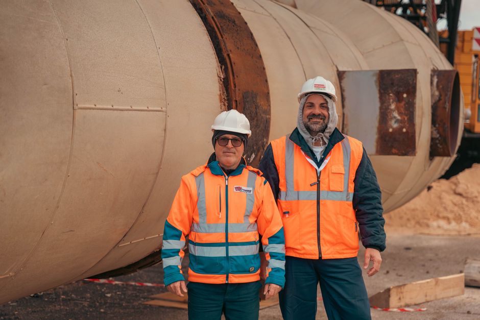 Two construction workers in safety gear standing by industrial machinery on a construction site.