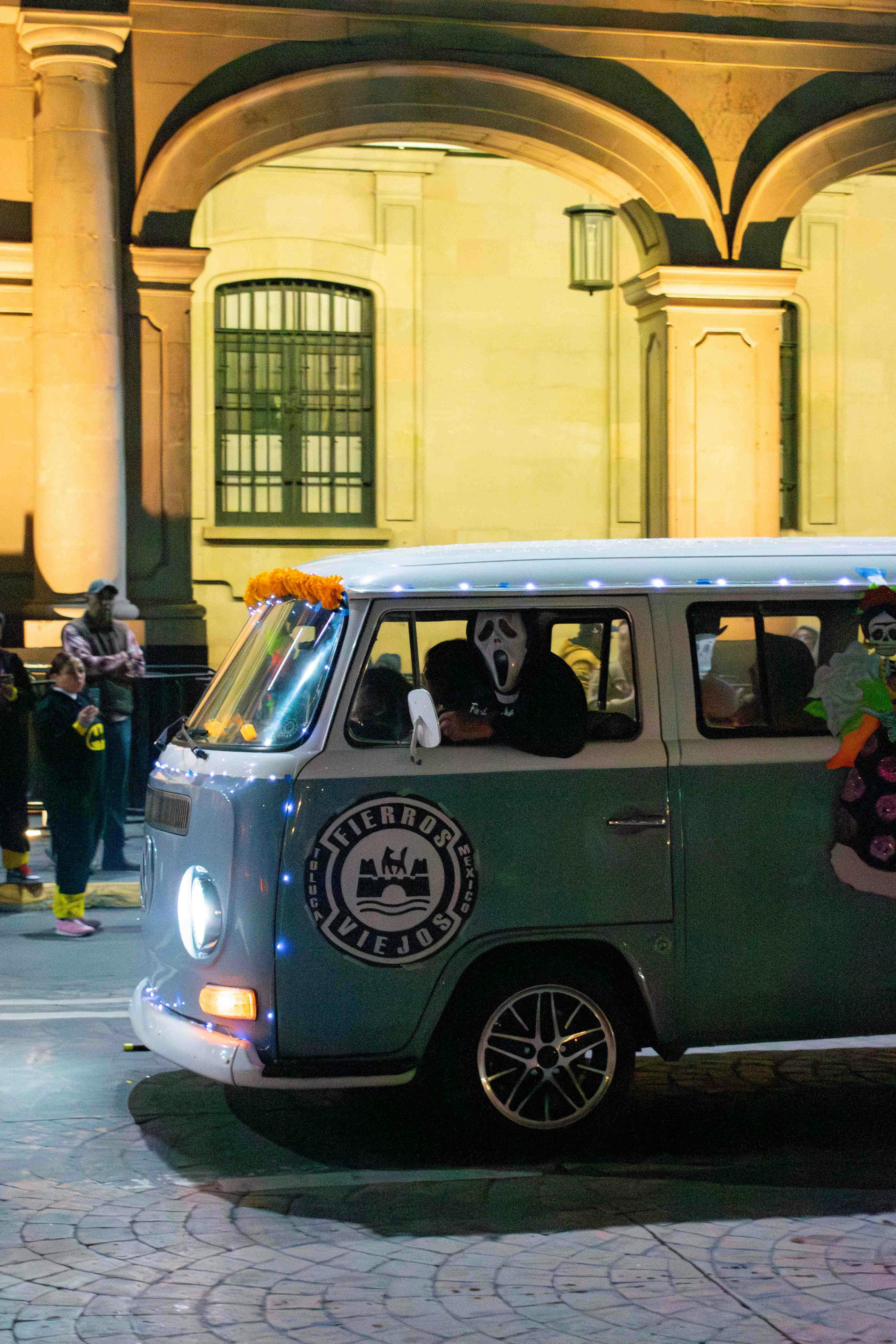 A vintage VW van decorated for Día de Muertos, captured at night during a lively parade.