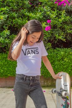 A cheerful young girl in a white t-shirt enjoying a sunny day with her skateboard outdoors.