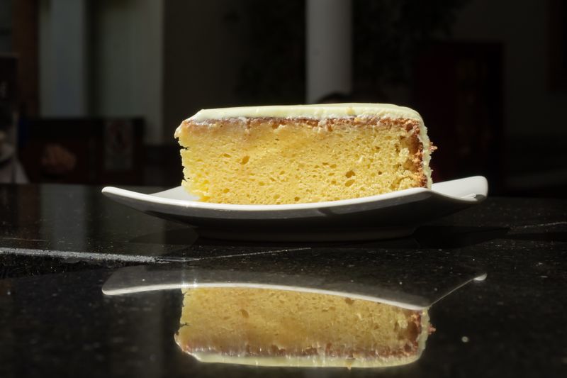 A fresh slice of lemon cake on a white plate, reflecting on a shiny black surface in a dimly lit café.