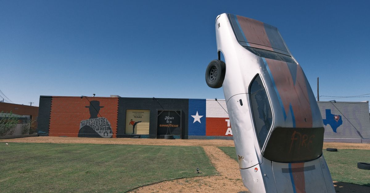 Art installation featuring a vertically planted car against Texas-themed murals under a clear blue sky.