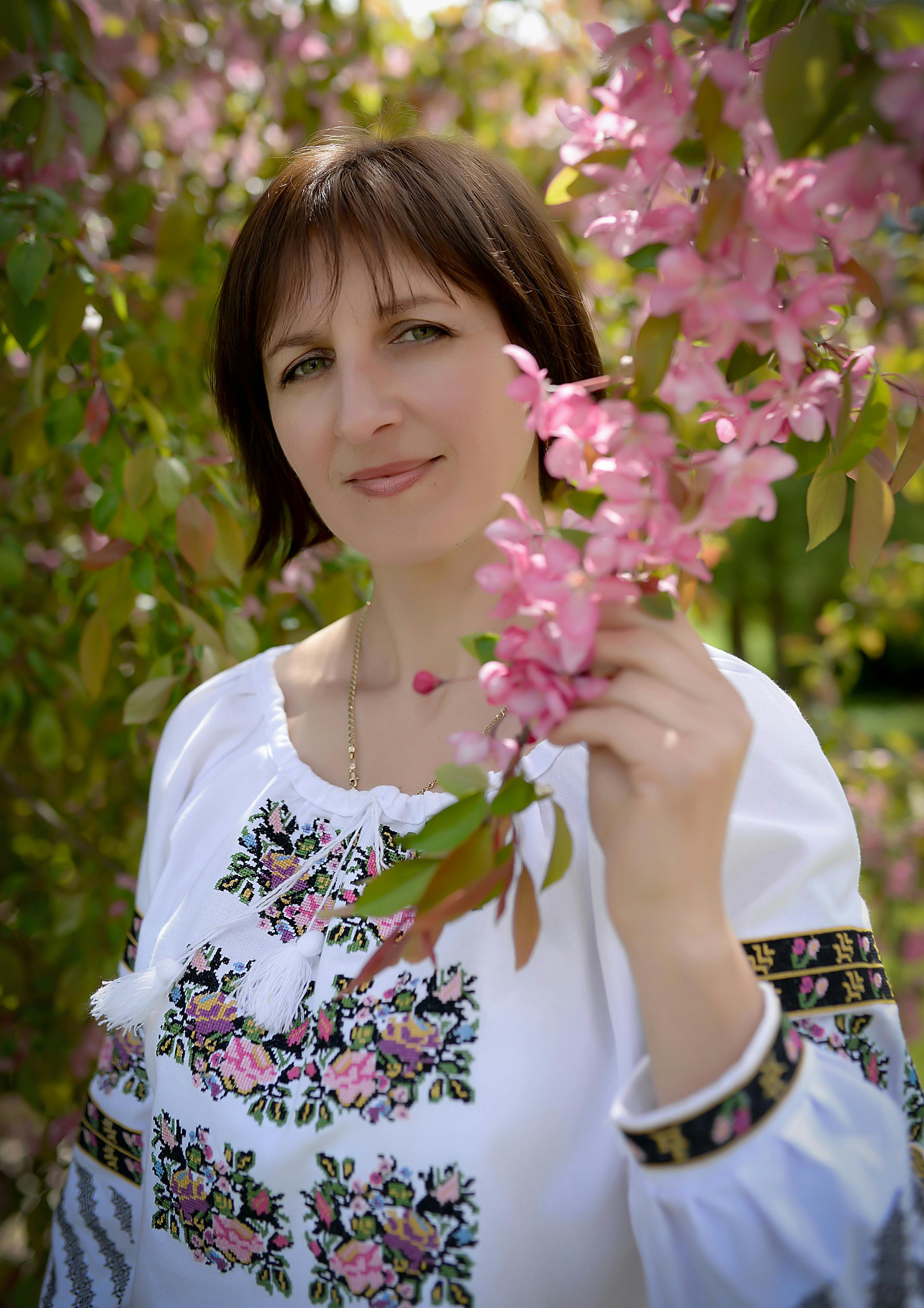 Woman in traditional embroidered dress holding pink blossoms in a garden.