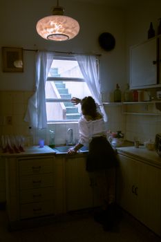 A woman gazes out a kitchen window as twilight casts a serene glow.