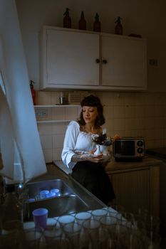 A vintage styled woman holding a potted plant stands in a dimly lit, cozy kitchen.