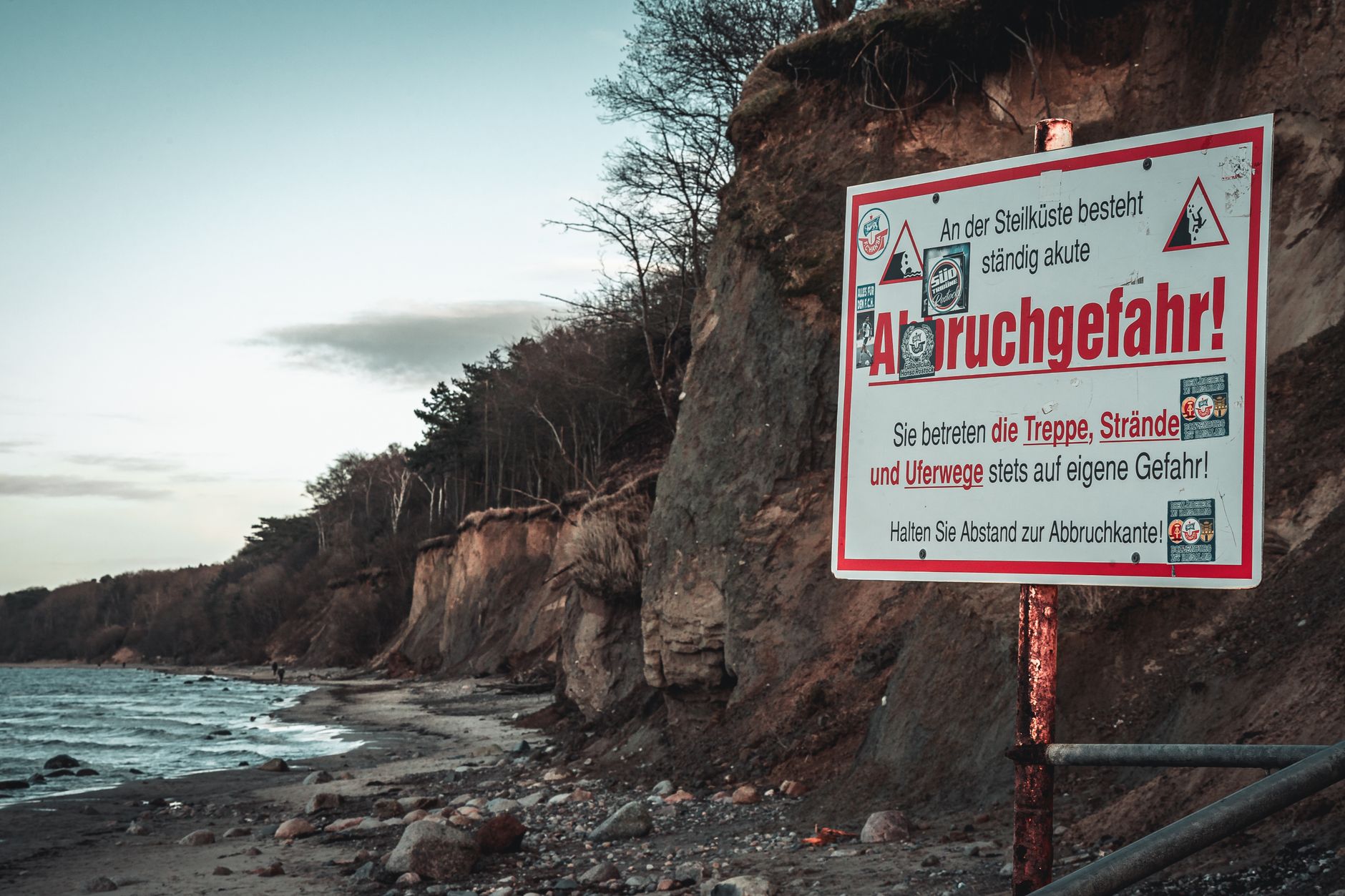 Warning sign on German coastline warns of erosion danger. Cliffside view at dusk with muted tones.