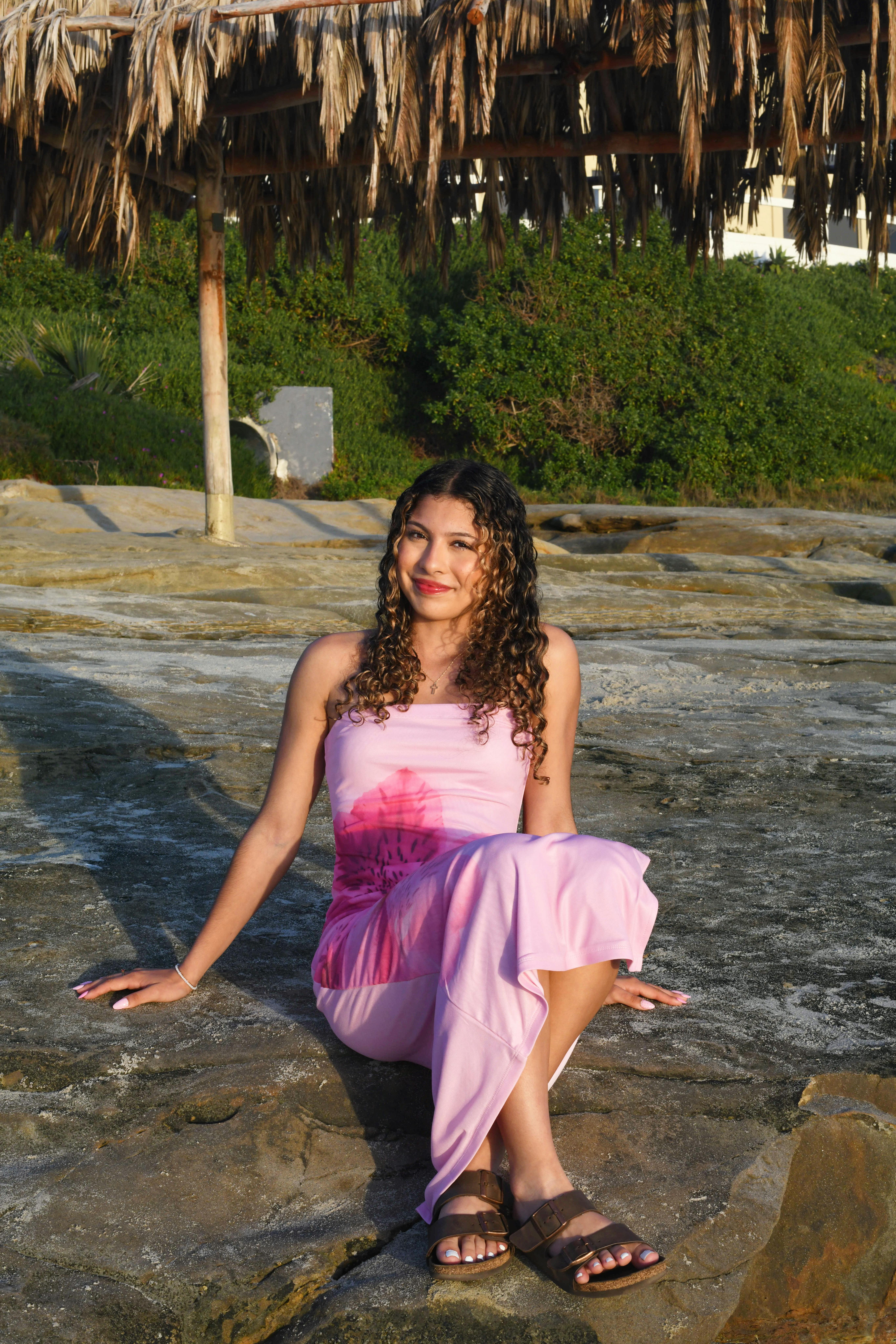 Smiling woman in a pink dress sitting on rocks under a thatched roof structure outdoors.