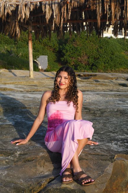 Smiling woman in a pink dress sitting on rocks under a thatched roof structure outdoors.